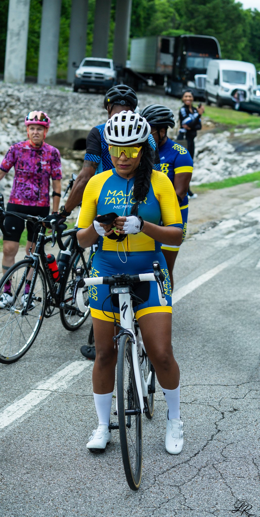 A woman in a yellow and blue cycling outfit, wearing a helmet and sunglasses, stands with her bicycle on the side of the road while looking at her phone. Other cyclists and vehicles are visible in the background.