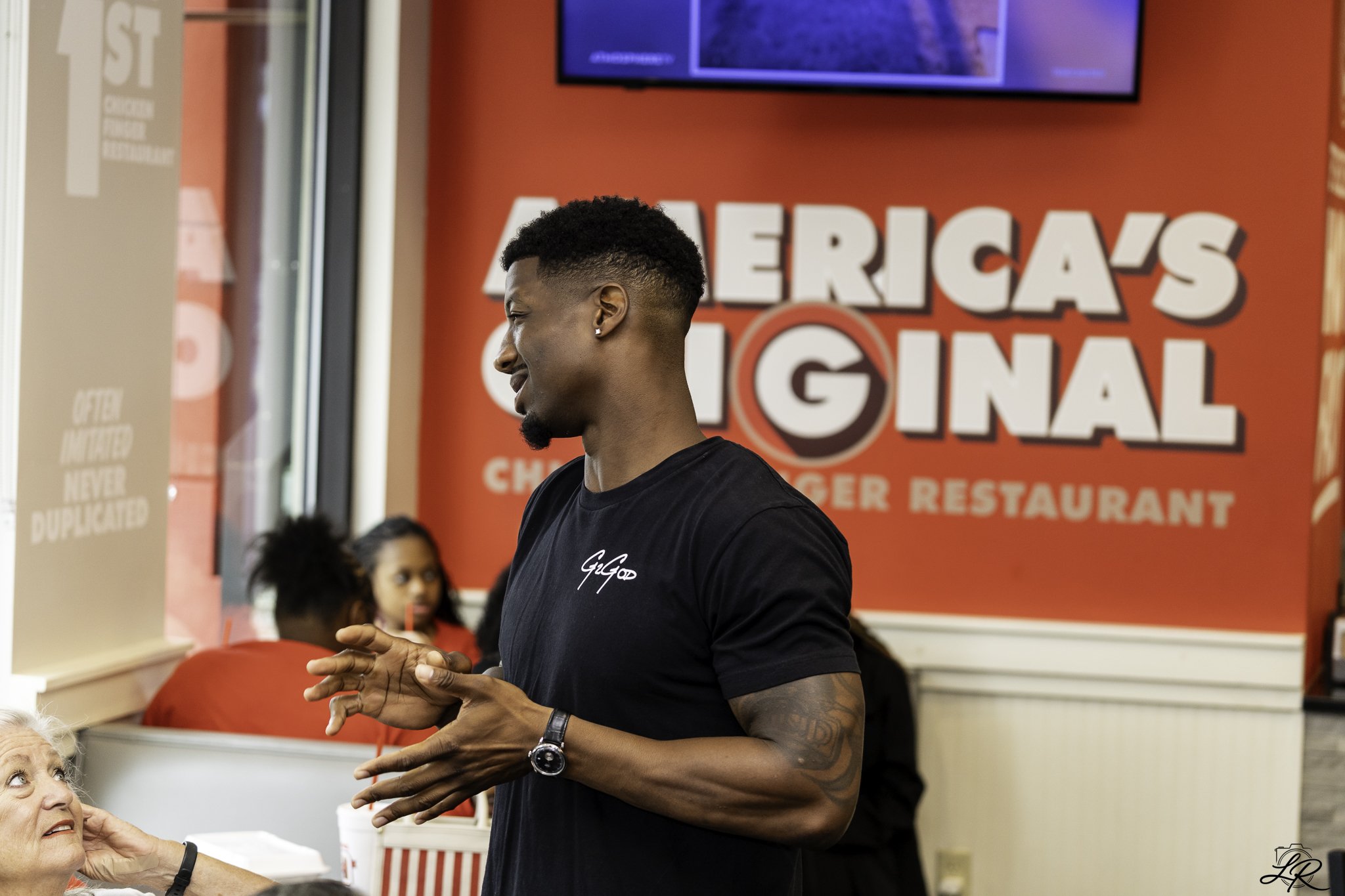 A young man with short curly hair, wearing a black t-shirt and wristwatch, is standing and talking to an elderly woman with white hair, who is looking up at him. They are inside a restaurant with red walls, featuring a sign that says 'American's Choi