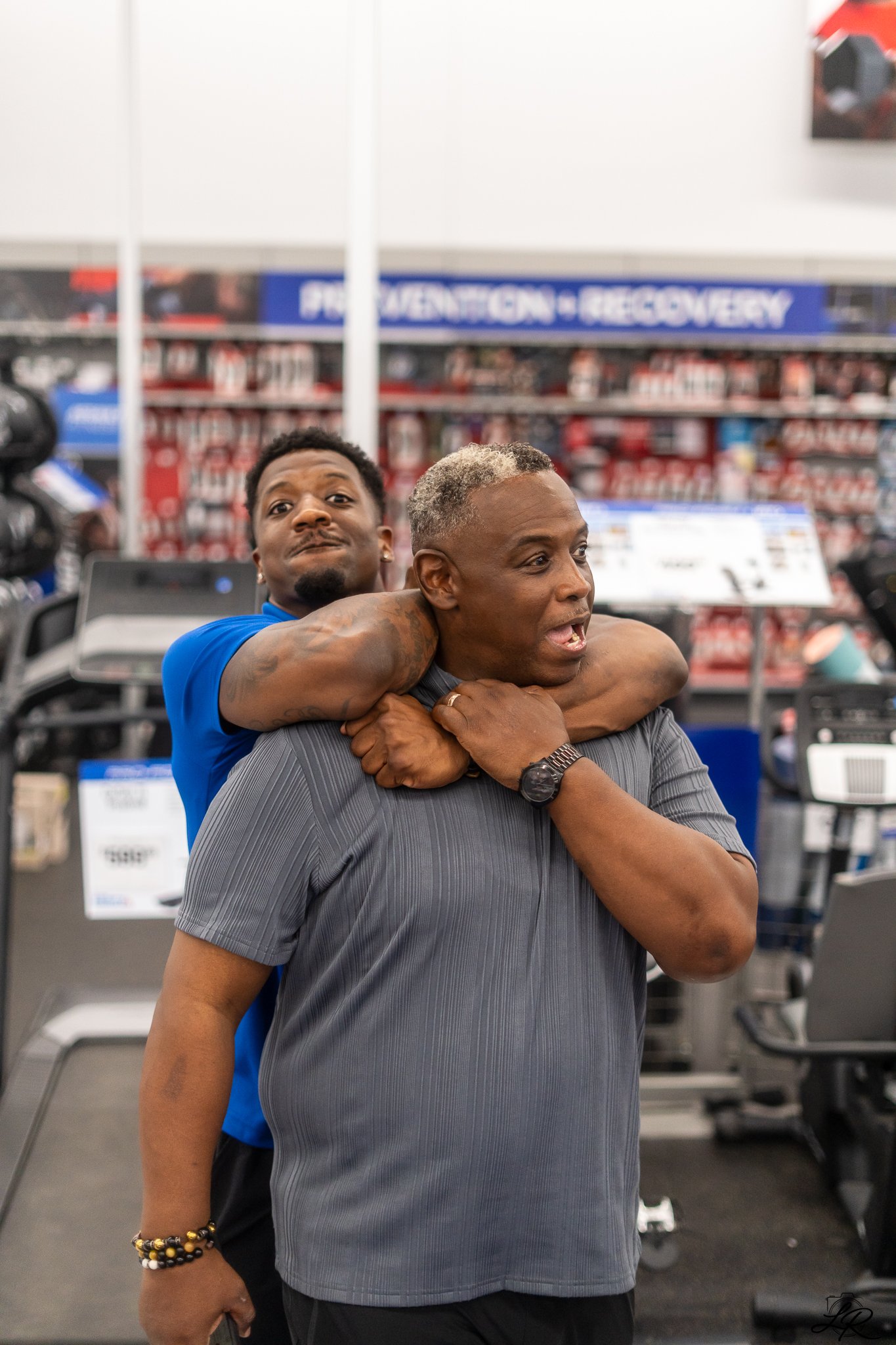 Two men embrace in a gym, with workout equipment and a banner reading 'PERFECTION - RECREVERY' in the background.
