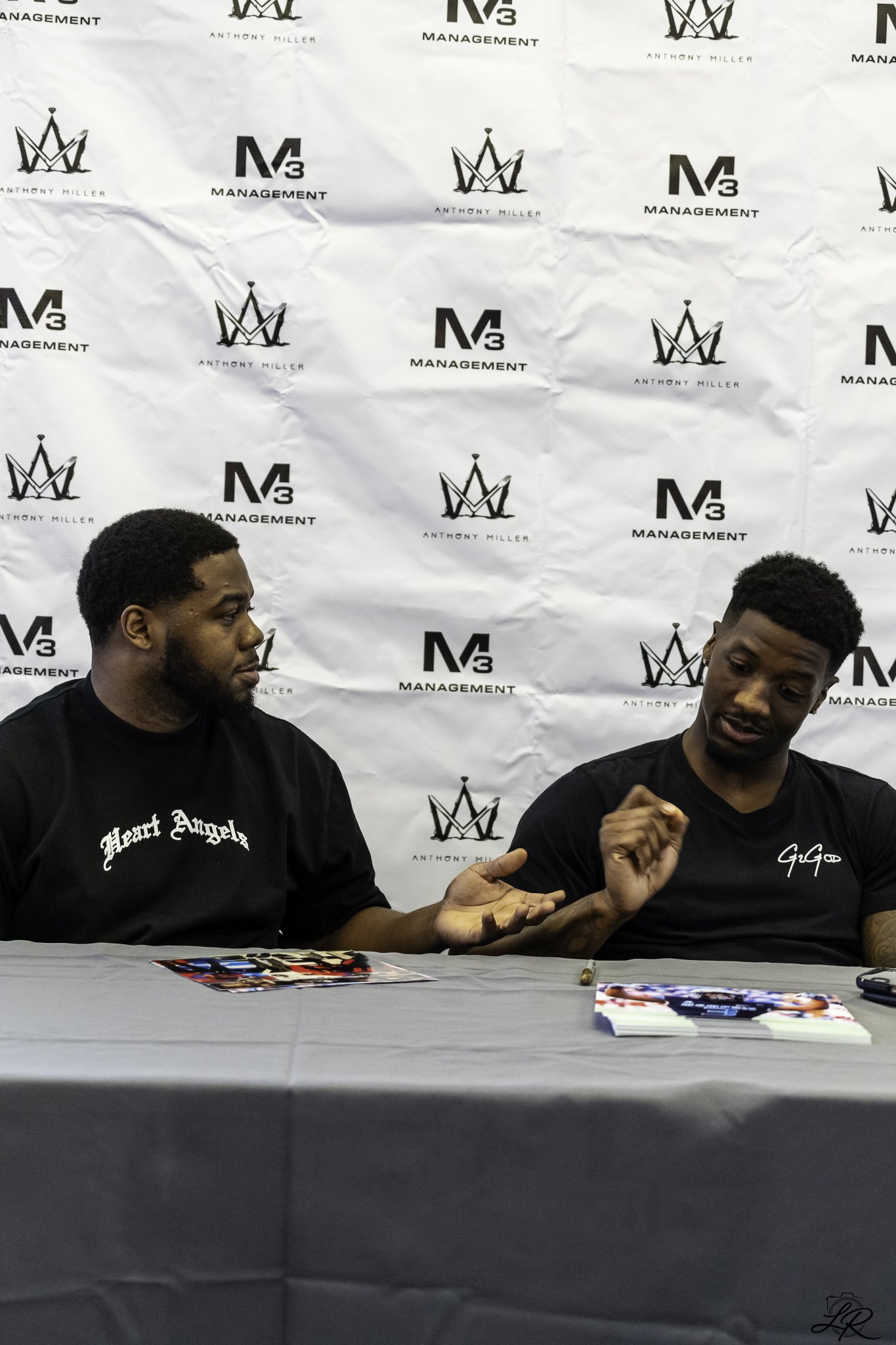 Two young men sitting at a table during an event, with a backdrop featuring logos that read 'Anthony Miller' and 'M3 Management.' They are engaged in conversation or signing autographs, with photographs and a pen on the table.