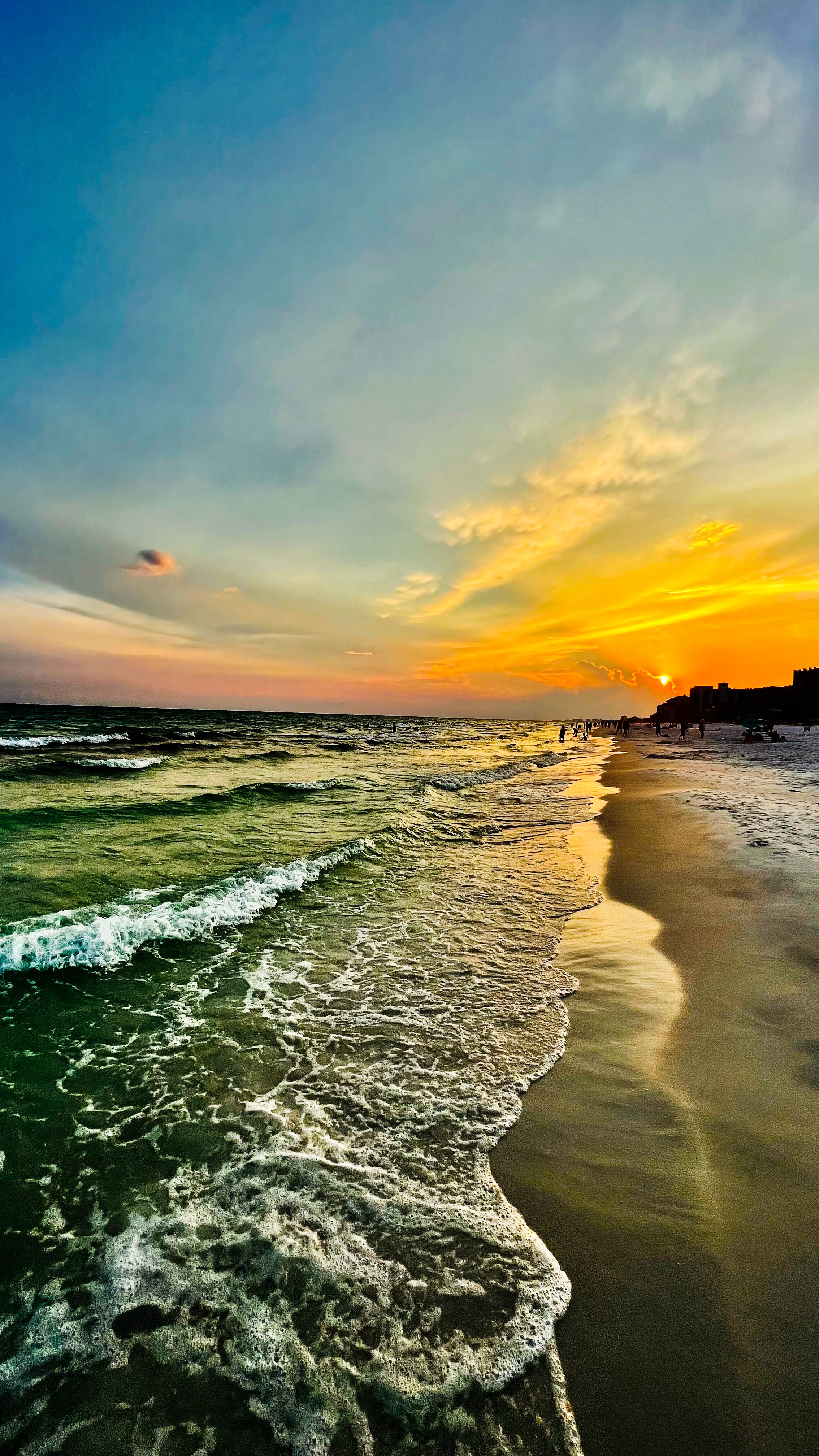 Sunset over the ocean with waves crashing on the sandy beach, people walking along the shoreline, and buildings in the distance.