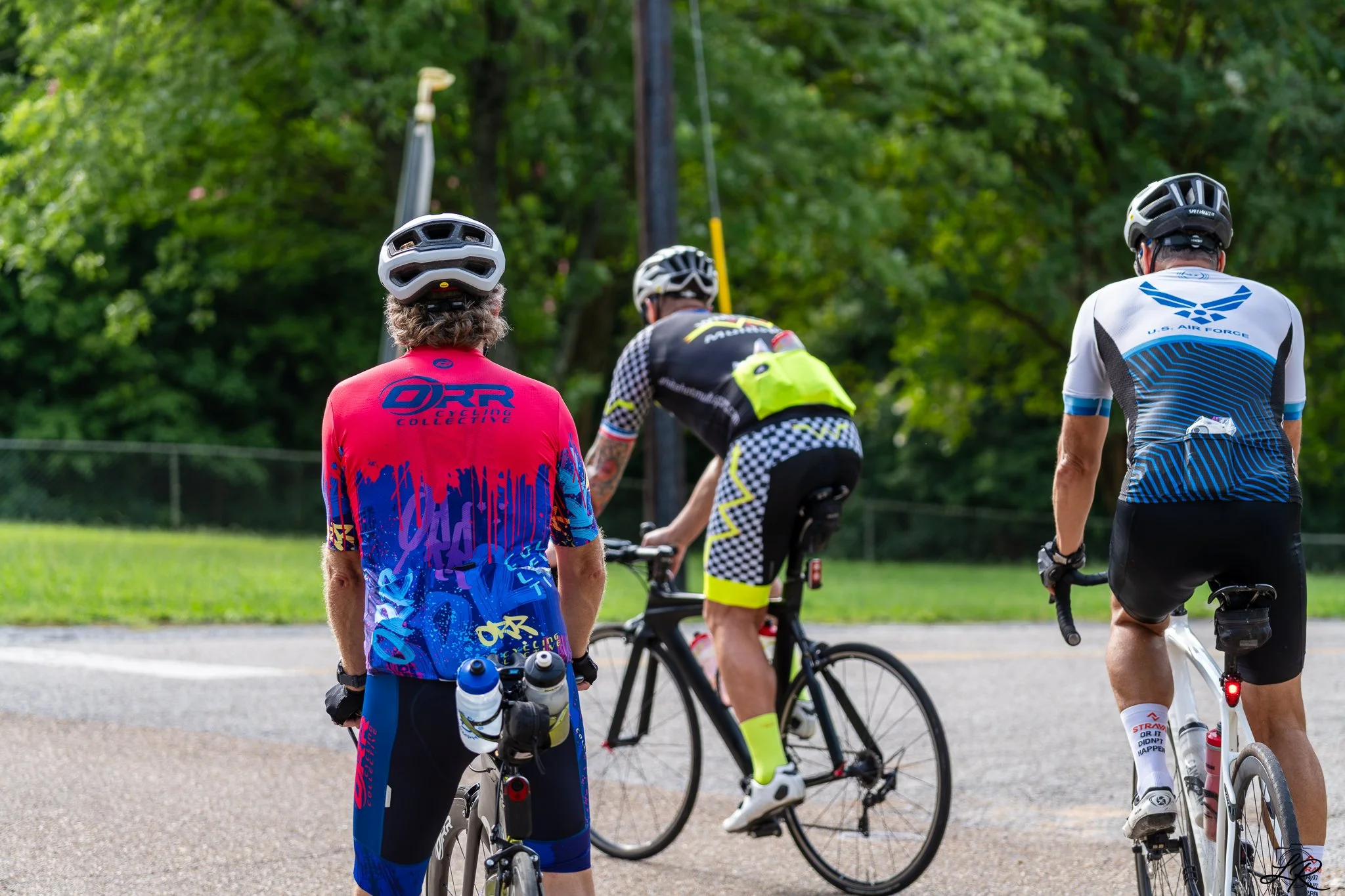 Group of cyclists wearing helmets and athletic gear, gathered outdoors on a paved path with green trees in the background.