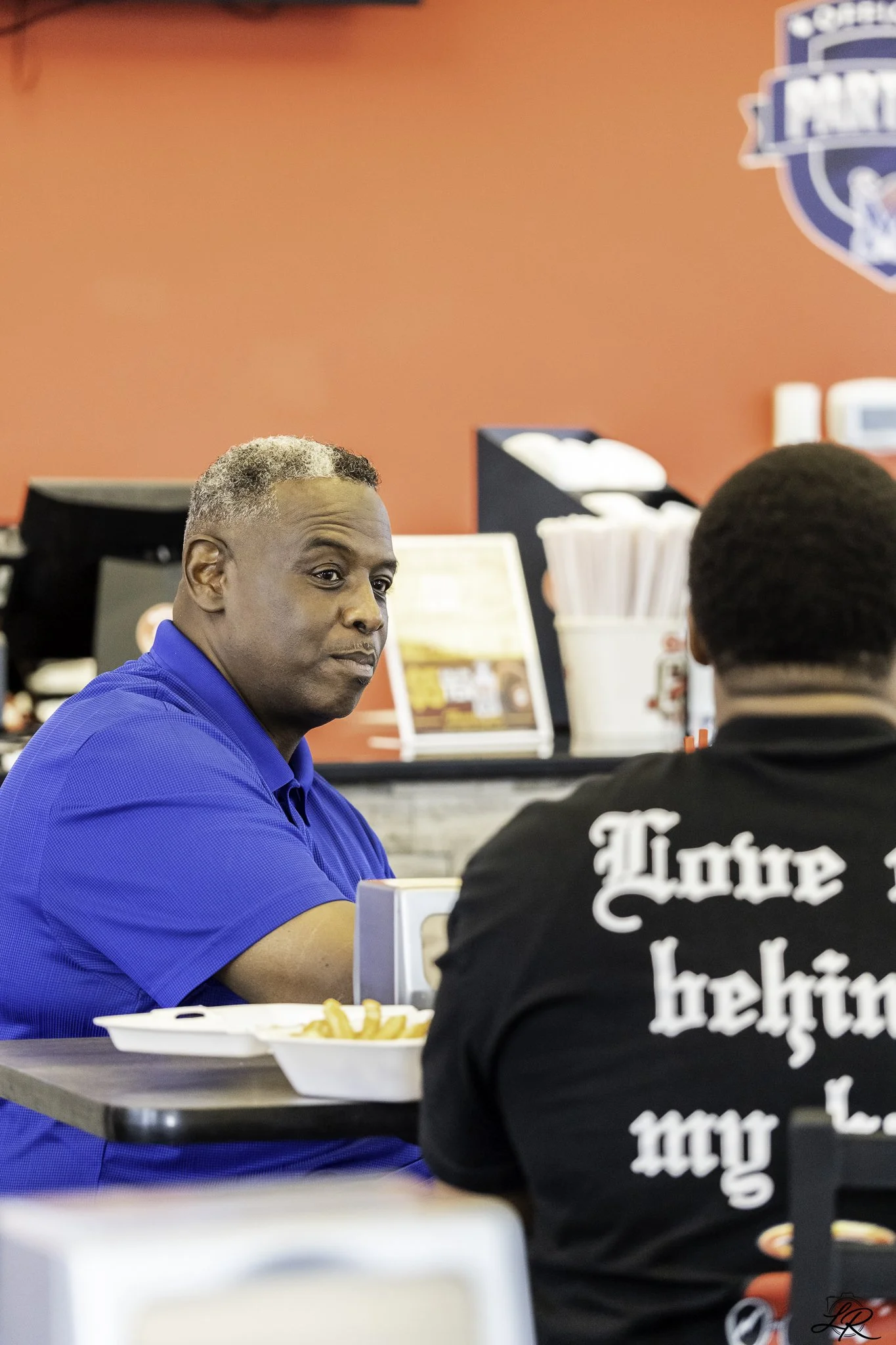 A middle-aged man with gray hair and wearing a blue polo shirt is sitting at a table in a restaurant, looking at a younger man with dark hair who is wearing a black shirt with white text on the back. There are fries in a white bowl on the table. The 