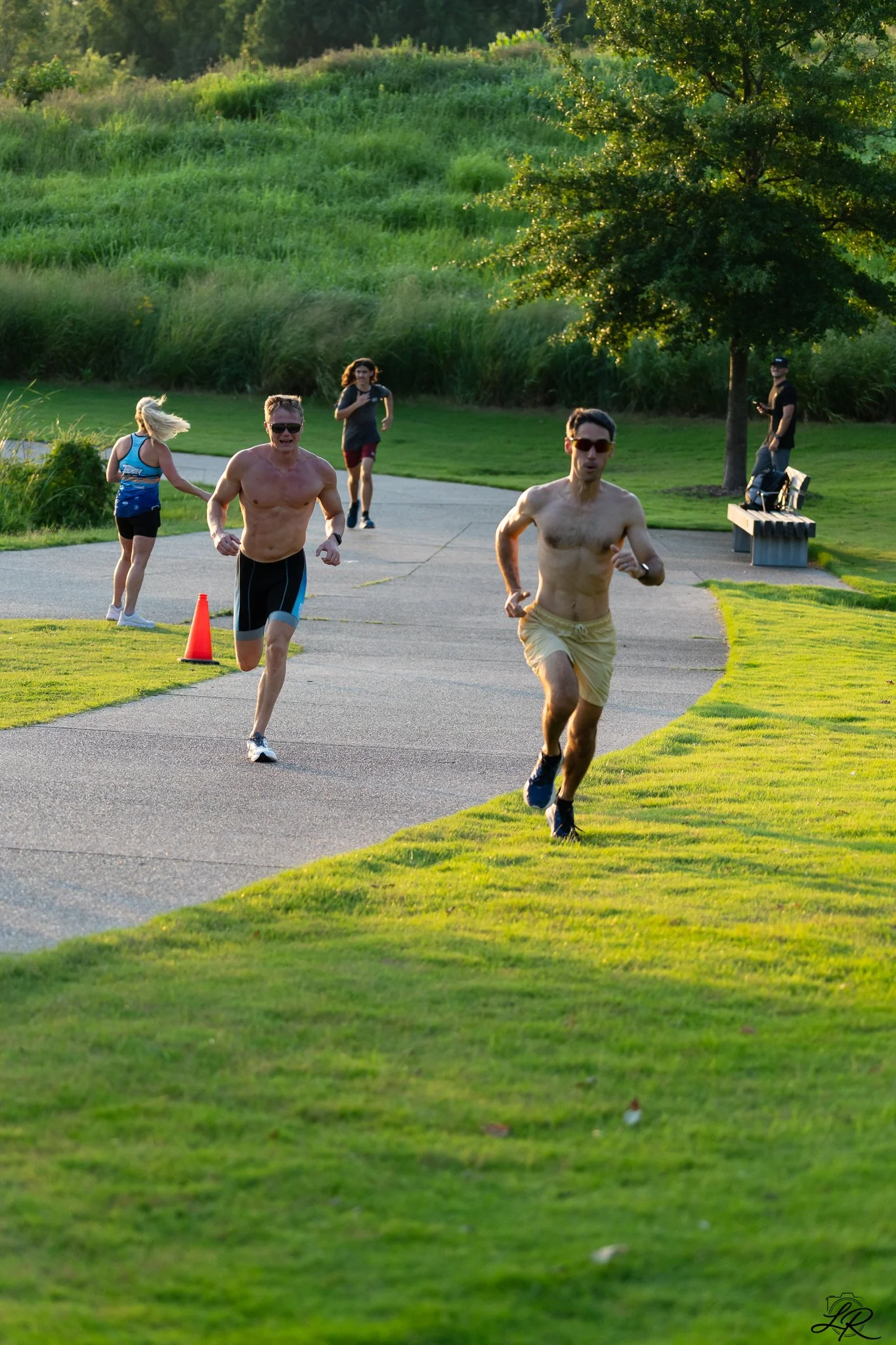 Two shirtless men and a woman running along a paved trail in a park with grassy hills and trees in the background. The men are wearing sunglasses, and one of the women is wearing a blue athletic top.