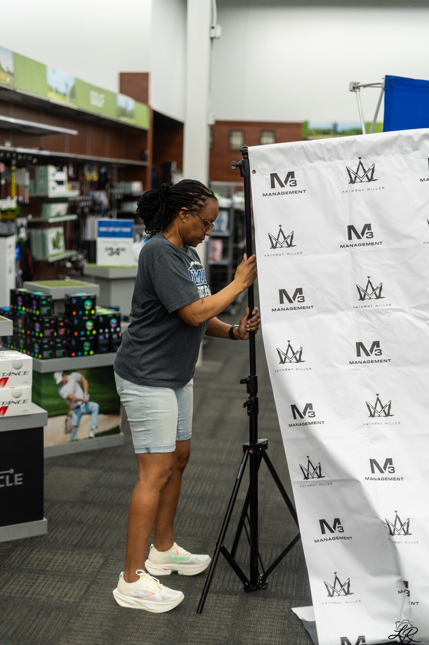 A woman with braided hair, wearing glasses, a gray t-shirt, light shorts, and white sneakers, is adjusting a white backdrop with the logos of N3 Management and Anthony Miller, inside a store with shelves of products in the background.