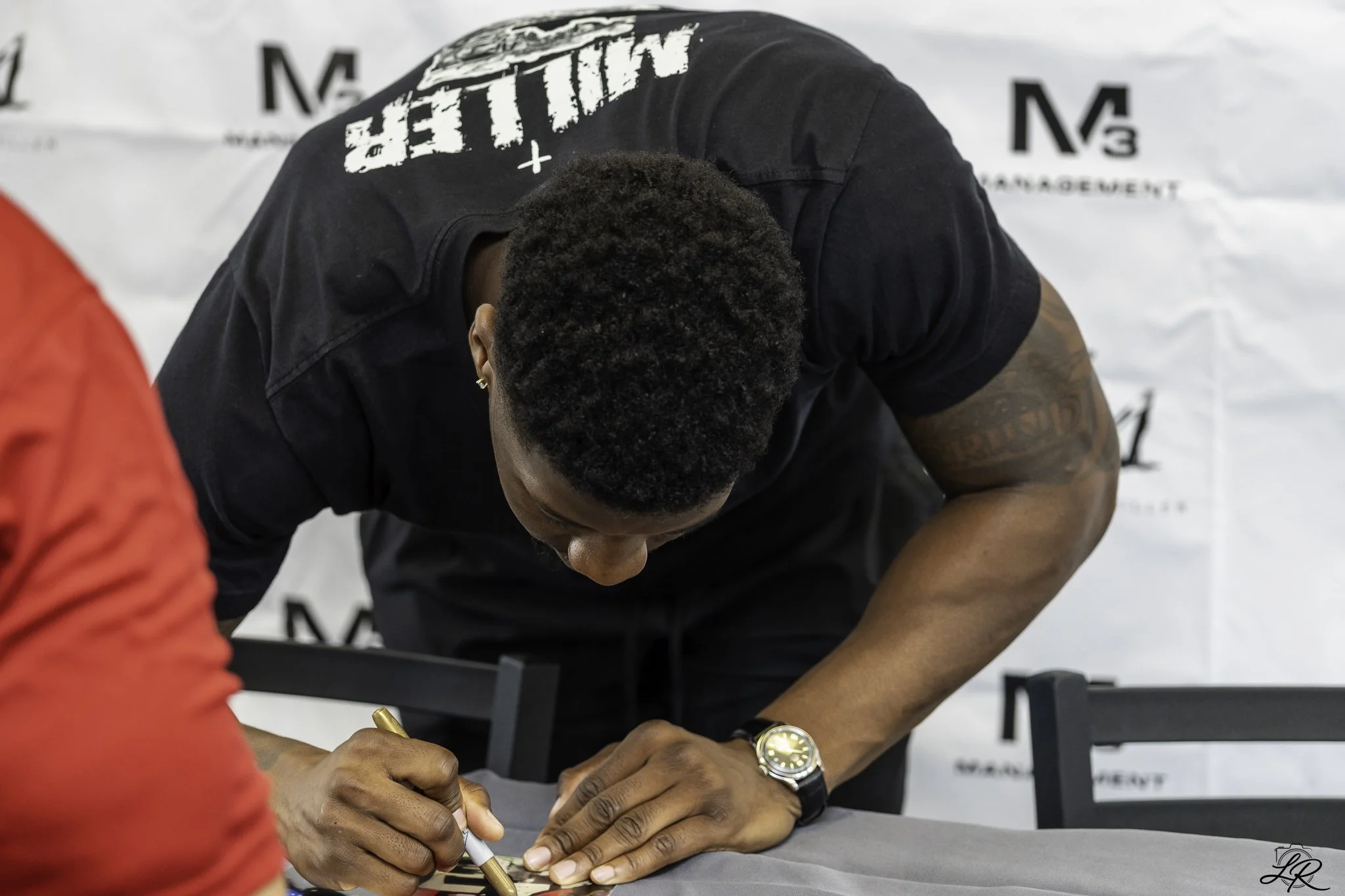 A man signing an autograph at a public event, wearing a black shirt and wristwatch, with a woman in red in the foreground.