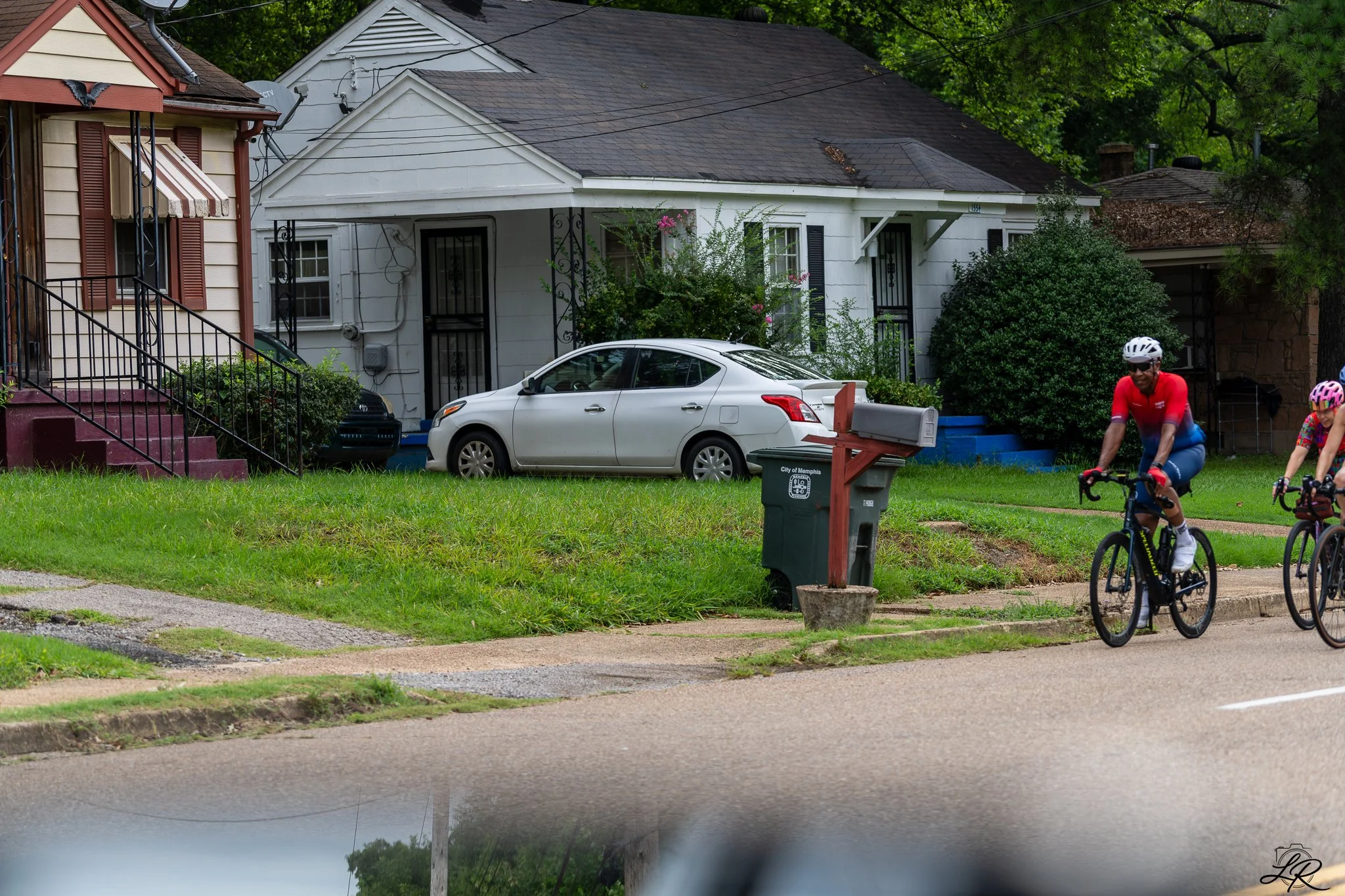A residential neighborhood with houses, a white car parked in front of a house, two people riding bicycles on the sidewalk, and lush green trees in the background.