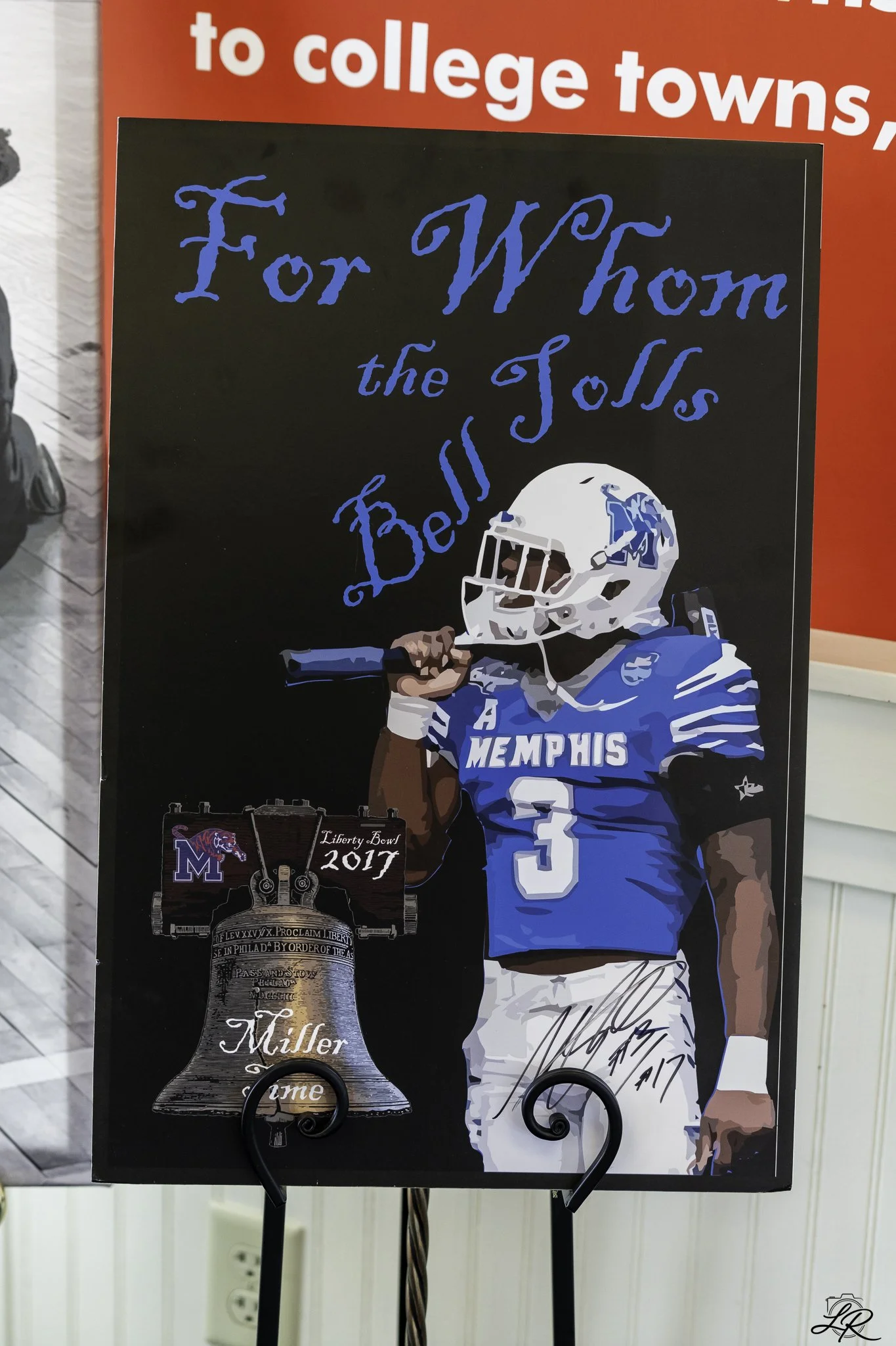 A memorial sign honoring a football player for Memphisis with a helmet and a football. The text reads, "For W Mom the Toss Beliss," with a small image of a football trophy with the words "Liberty Bowl 2017." The sign has red at the top and is attache