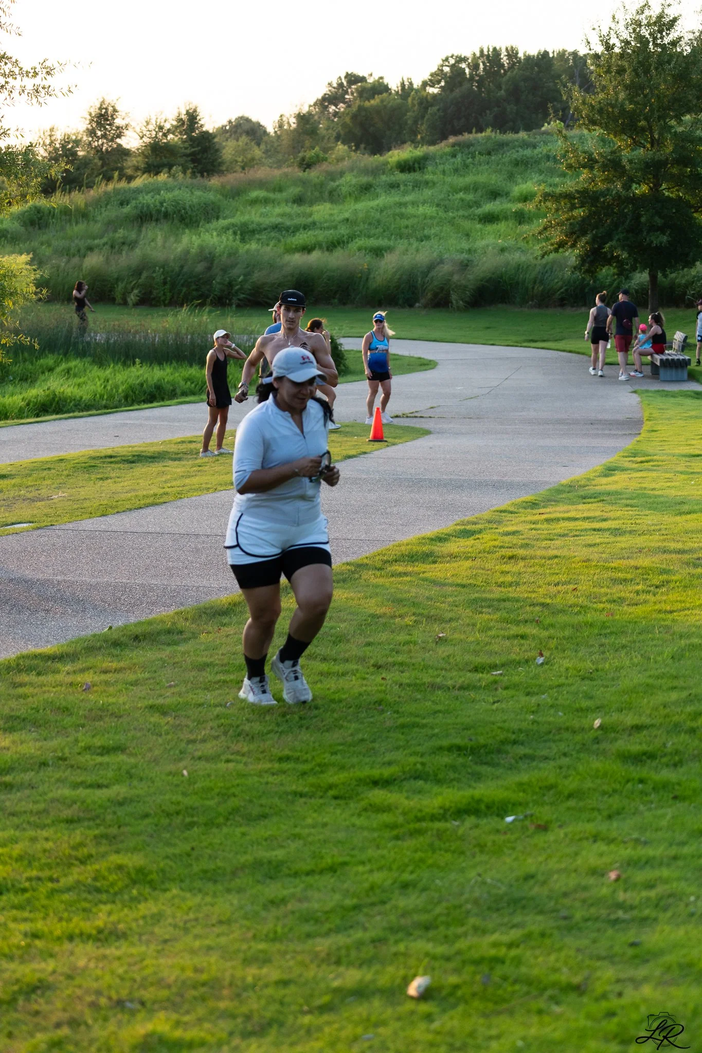 A group of people running and walking on a paved pathway in a park during late afternoon or early evening with green grass, trees, and a hillside in the background.