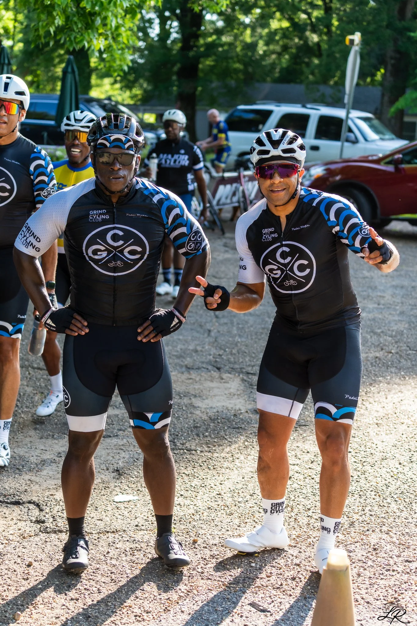 Group of cyclists in matching black, white, and blue jerseys standing outdoors on a gravel surface, with trees and parked cars in the background.