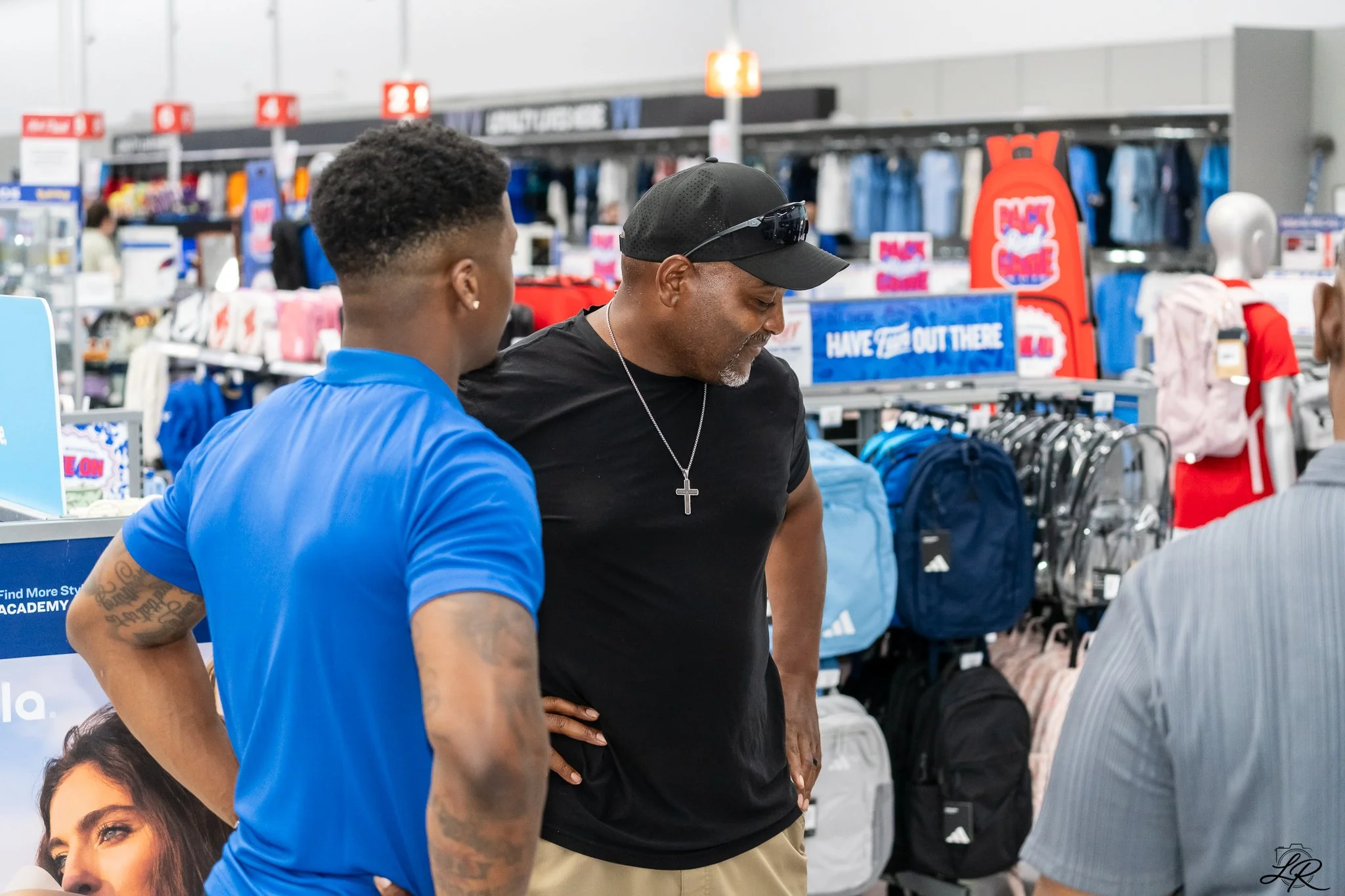 Two men standing and talking inside a retail store, with shelves of apparel and promotional signs in the background.