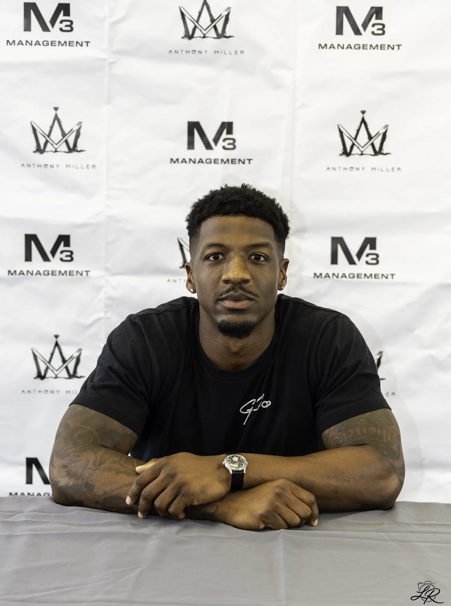 A young man with tattoos on his arms and a watch on his wrist, sitting at a table with his arms crossed. He is wearing a black shirt and looking directly at the camera. The background features a white banner with black logos and text, including 'N3 M