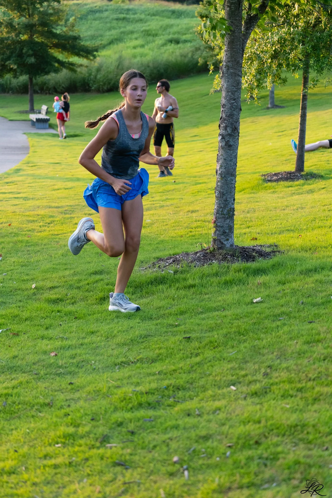 A young woman running on a grassy park, wearing a gray tank top, blue shorts, and gray sneakers, with trees and other park visitors in the background.