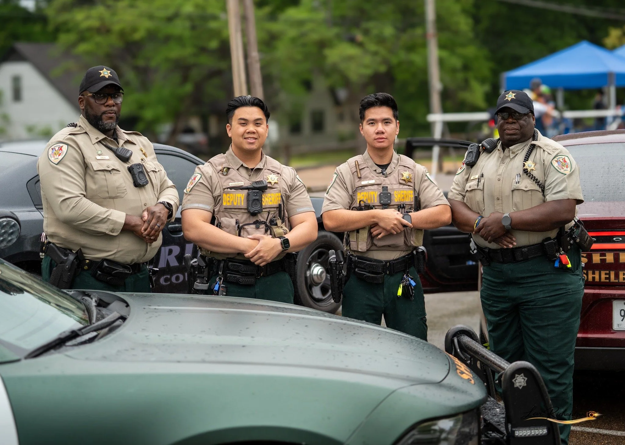 Four sheriff deputies standing in a row outdoors, wearing uniforms and gear, with cars and a blue tent in the background.