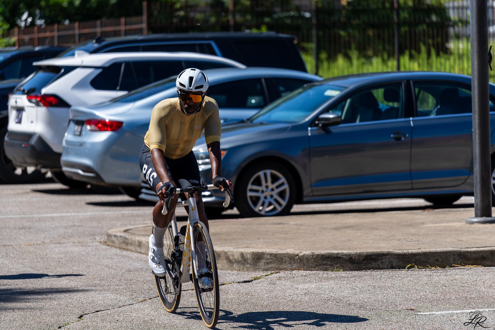 A cyclist wearing a helmet and sunglasses riding a bike in a parking lot with parked cars in the background.
