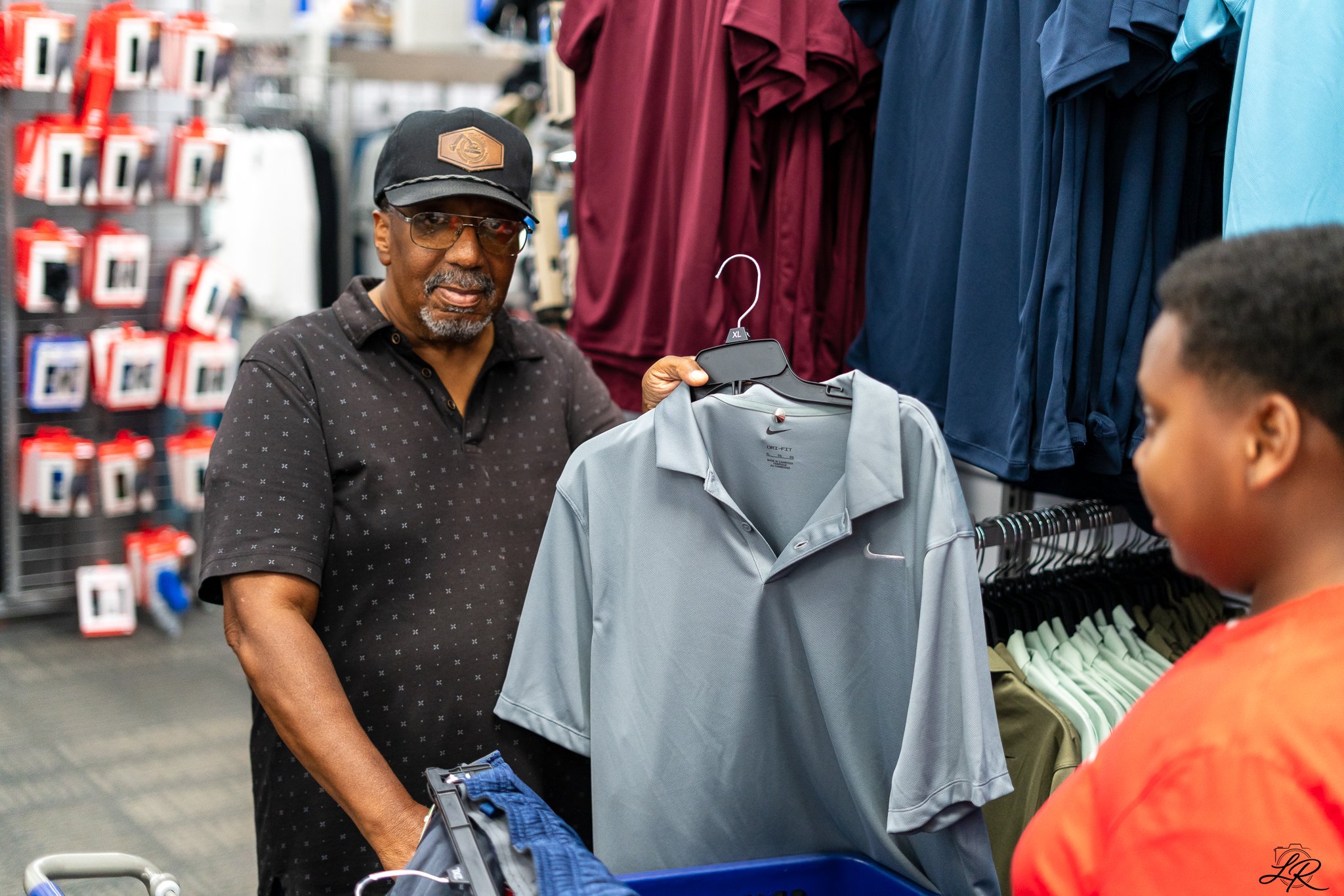 An older man wearing glasses, a black cap, and a black shirt with small white dots shopping with a young boy in an aisle of a store with clothing on racks and shelves.
