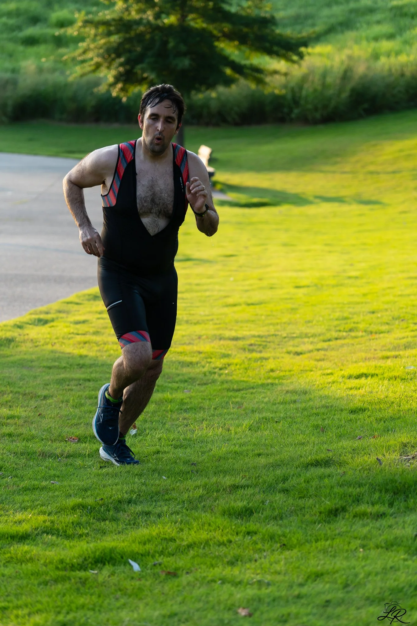 A man running outdoors on a grassy area during daylight, wearing a sleeveless running outfit with a backpack and athletic shoes.