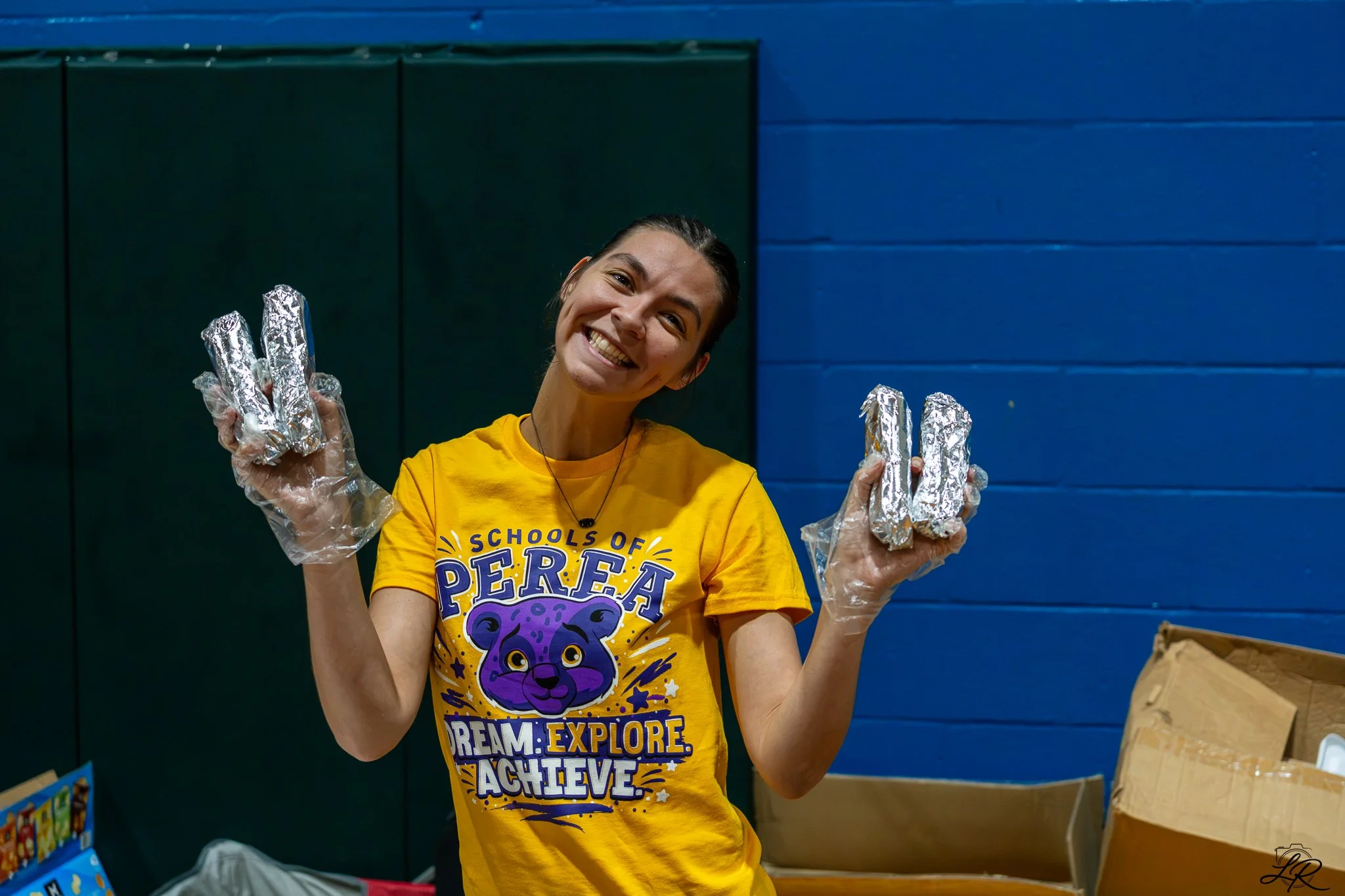 A young woman with a bright smile, wearing a yellow t-shirt with purple text and graphics, holds foil-wrapped items in both hands, standing in front of a blue and green wall.
