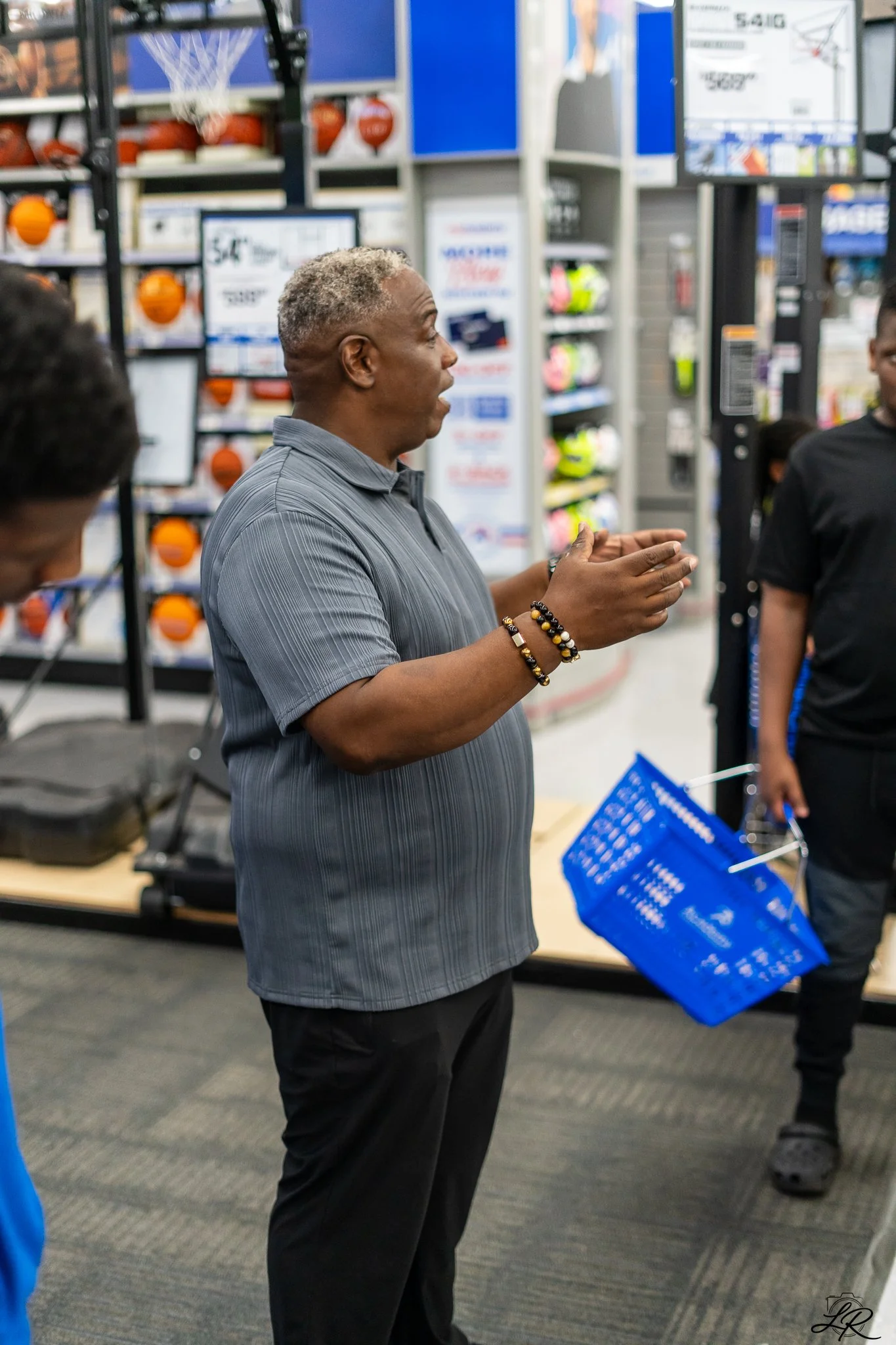 A man with gray hair wearing a striped gray shirt and black pants standing and talking, holding his hands together in an indoor sports store. Behind him are shelves with basketballs, helmets, and sports equipment, as well as monitors and signage.