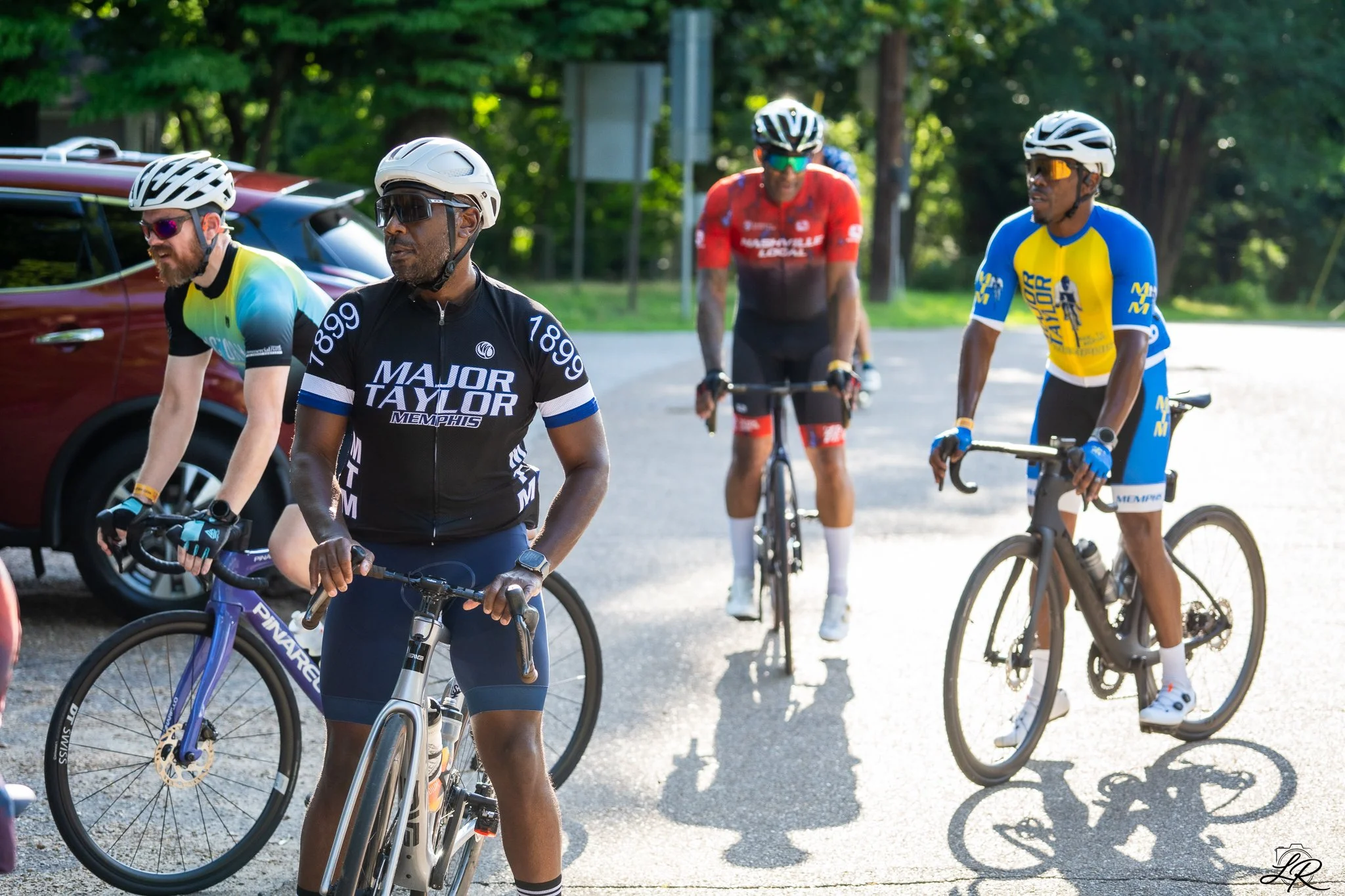 Four cyclists with helmets and sportswear riding bicycles on a paved road surrounded by trees.