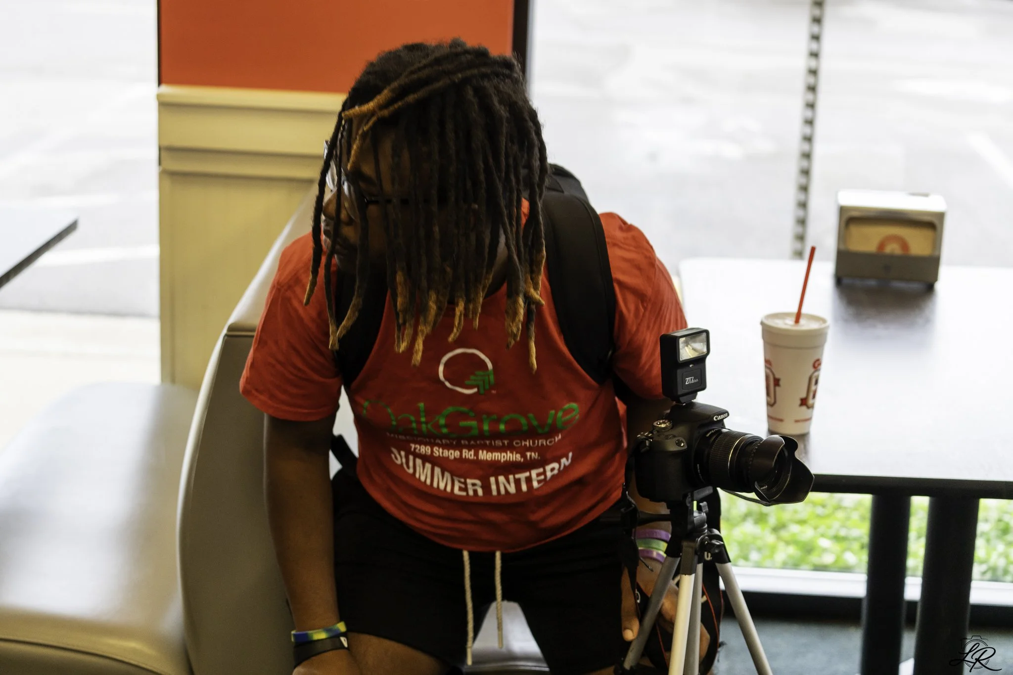 A person with dreadlocks wearing glasses and a red T-shirt with green text, standing behind a camera and a drink on a table, inside a building near a window.