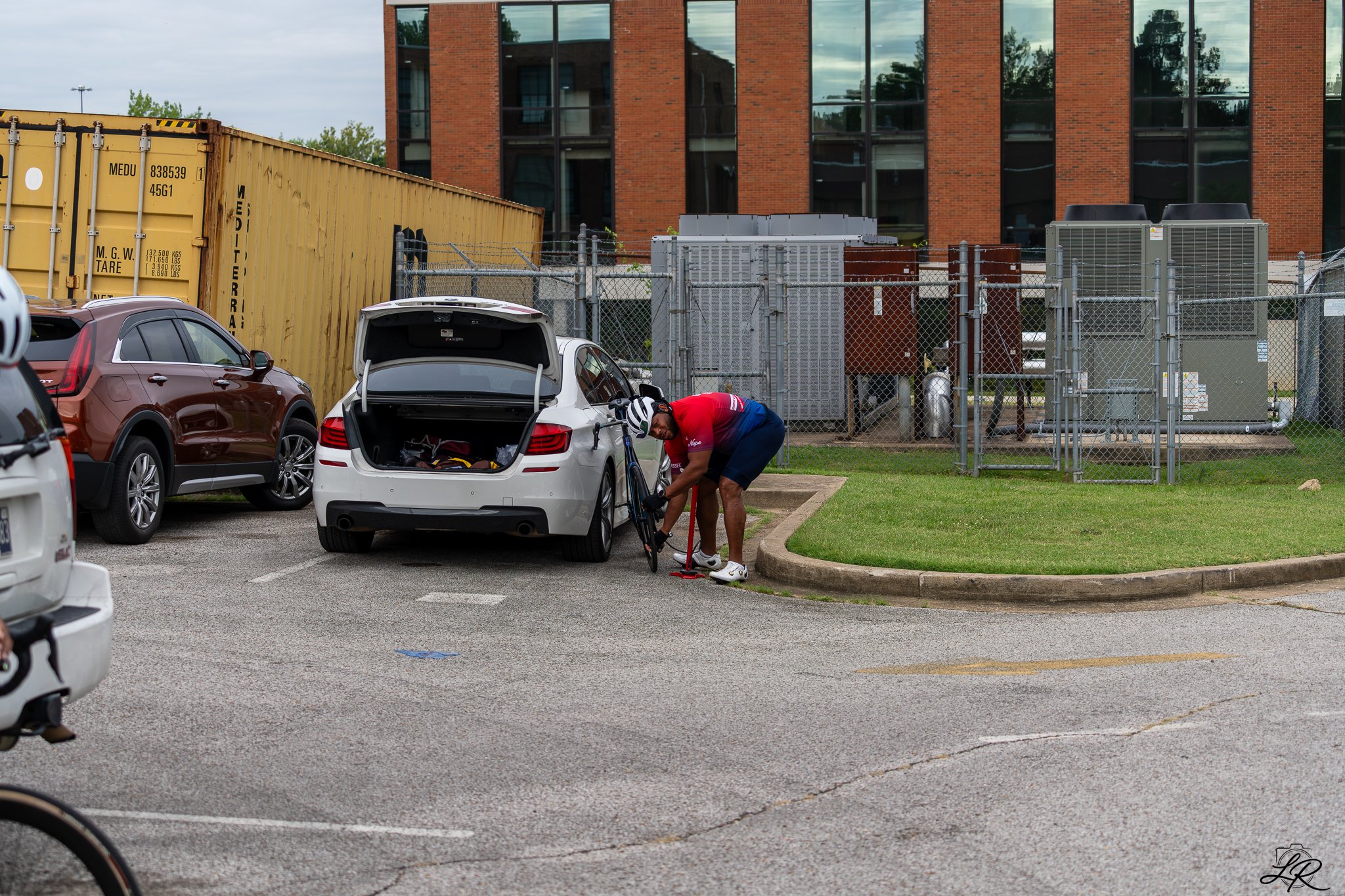 A man in a red shirt and blue shorts is fixing his bicycle near a white car with its trunk open in a parking lot. There are other vehicles and a brick building with large windows in the background, along with a chain-link fence and industrial equipme