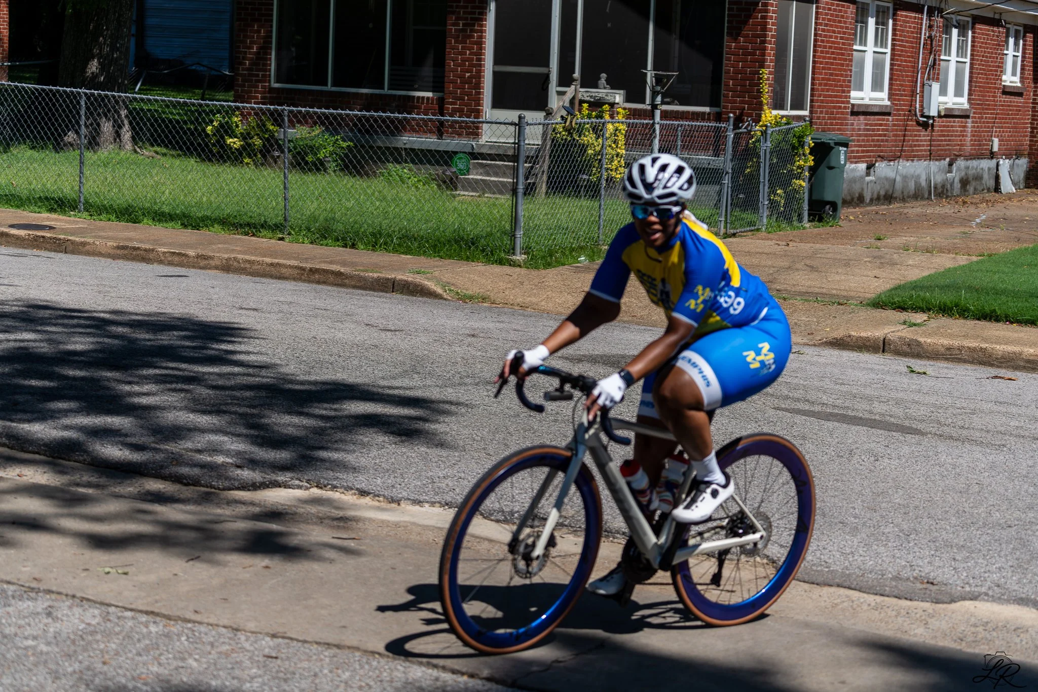 A person wearing a yellow and blue cycling jersey, black helmet, and sunglasses riding a bicycle on a residential street with a brick house and chain-link fence in the background.