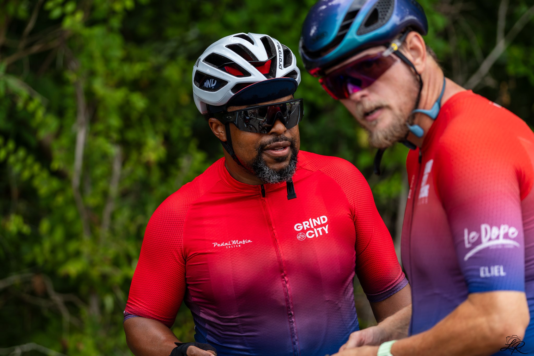 Two male cyclists wearing helmets and sports glasses having a conversation outdoors with green foliage in the background.