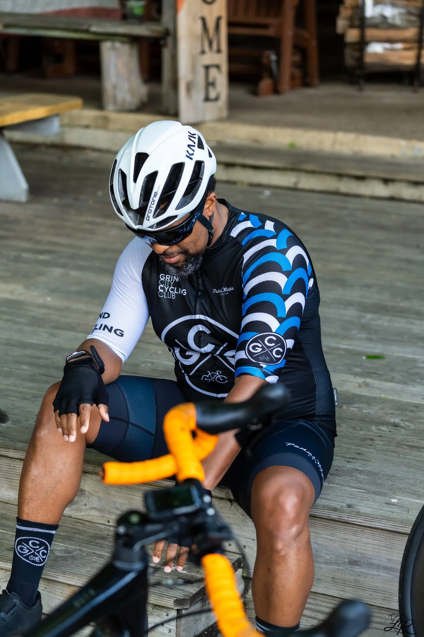 A man in cycling gear, including a white helmet and sunglasses, is sitting on wooden steps, adjusting his leg after riding a bike. The background features rustic wooden structures or furniture.