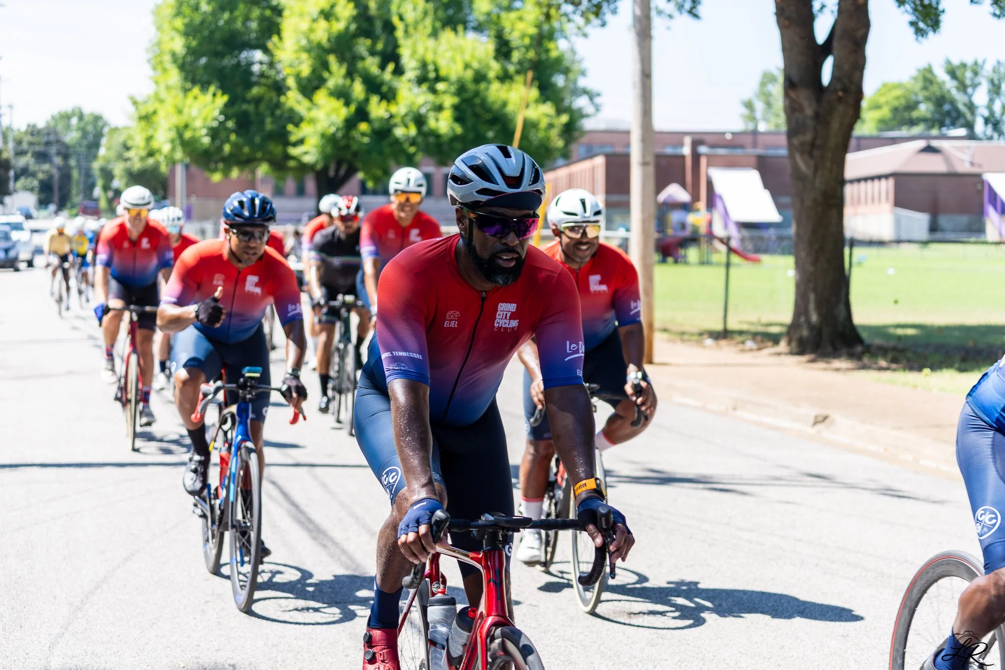 A group of cyclists riding together on a sunny day in a park or neighborhood, wearing helmets and colorful cycling gear.