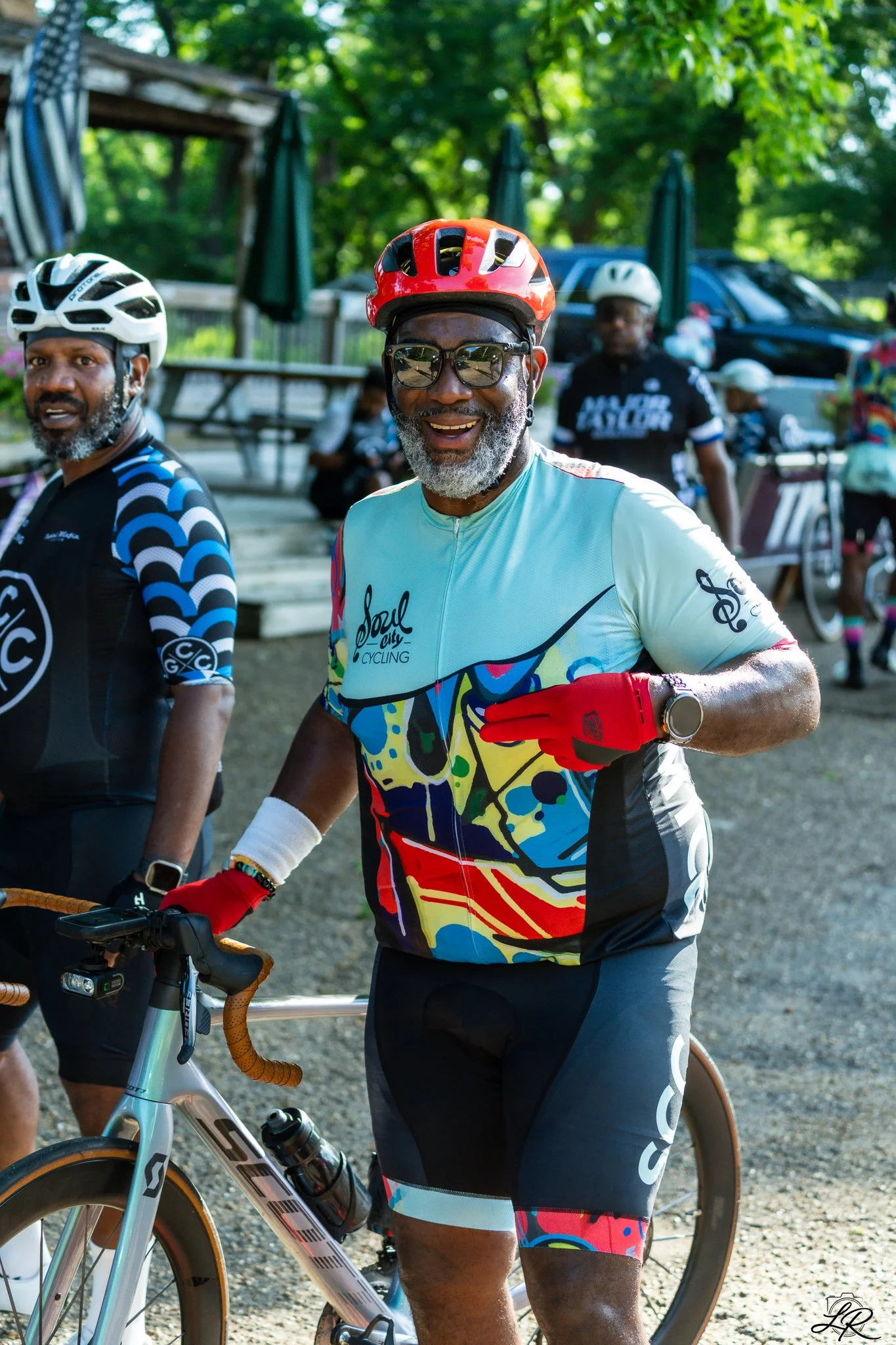 Group of older men in cycling gear with helmets, sunglasses, and colorful jerseys at an outdoor cycling event, with trees and cars in the background.