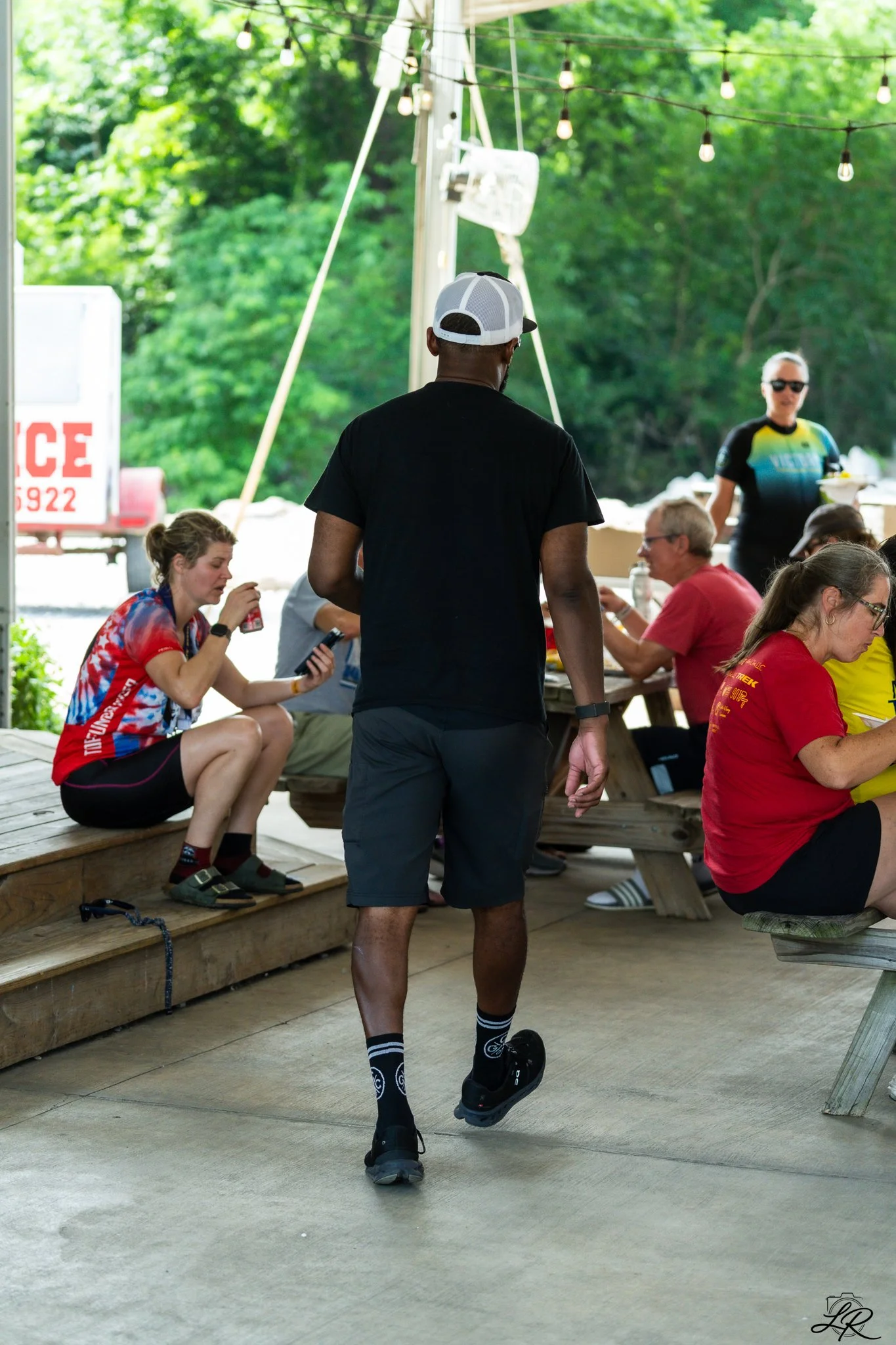 A man jogging in athletic gear with a black t-shirt, gray shorts, black sneakers, and a white cap is at a picnic table, while a group of people sitting at the table are watching him. The setting appears to be outdoors under string lights, with greene