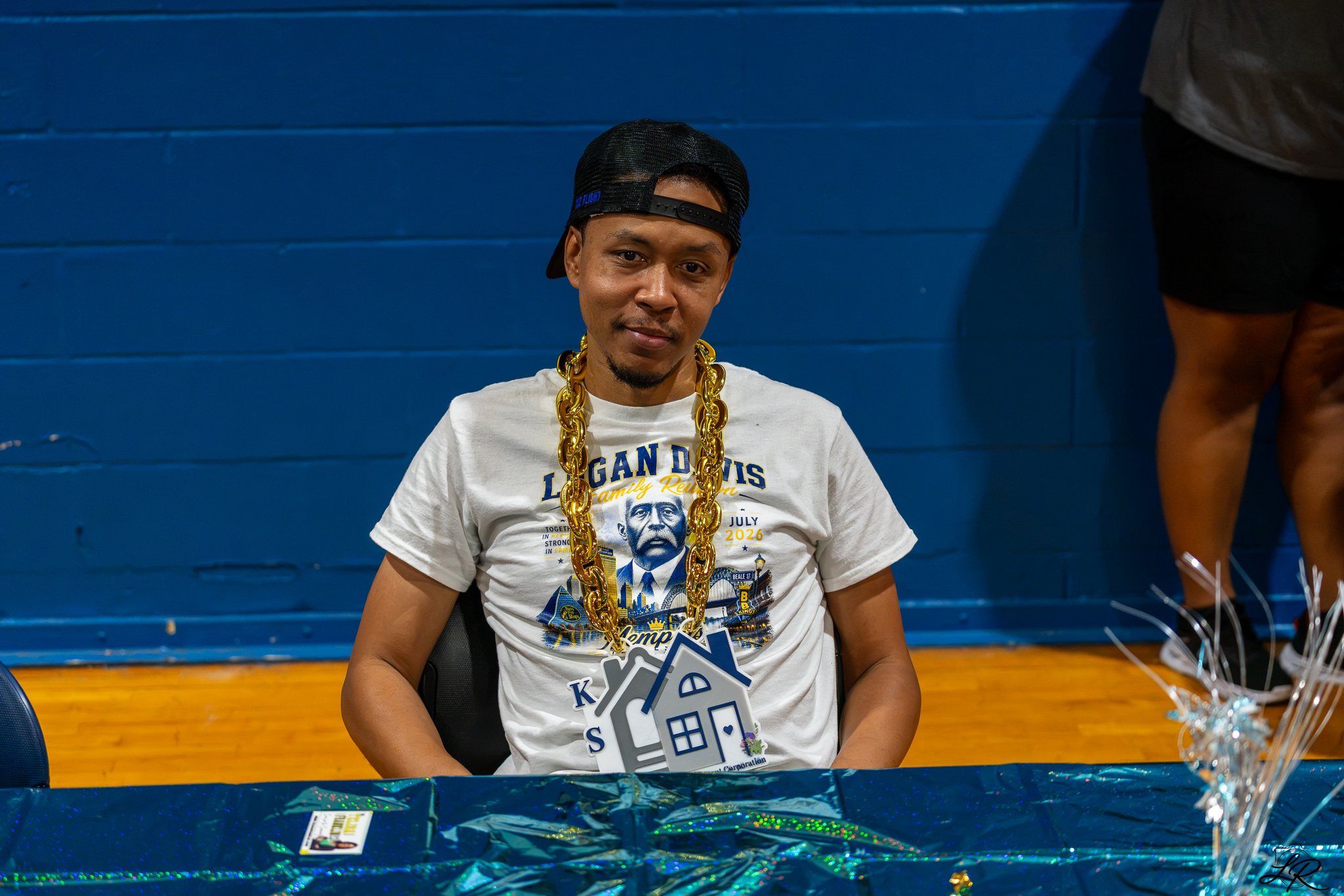A young man sitting at a table, wearing a black cap backwards, a white T-shirt with a graphic of a person and the words 'Louisiana family reunion,' and a large gold chain necklace. He is in a room with a blue painted brick wall in the background, and