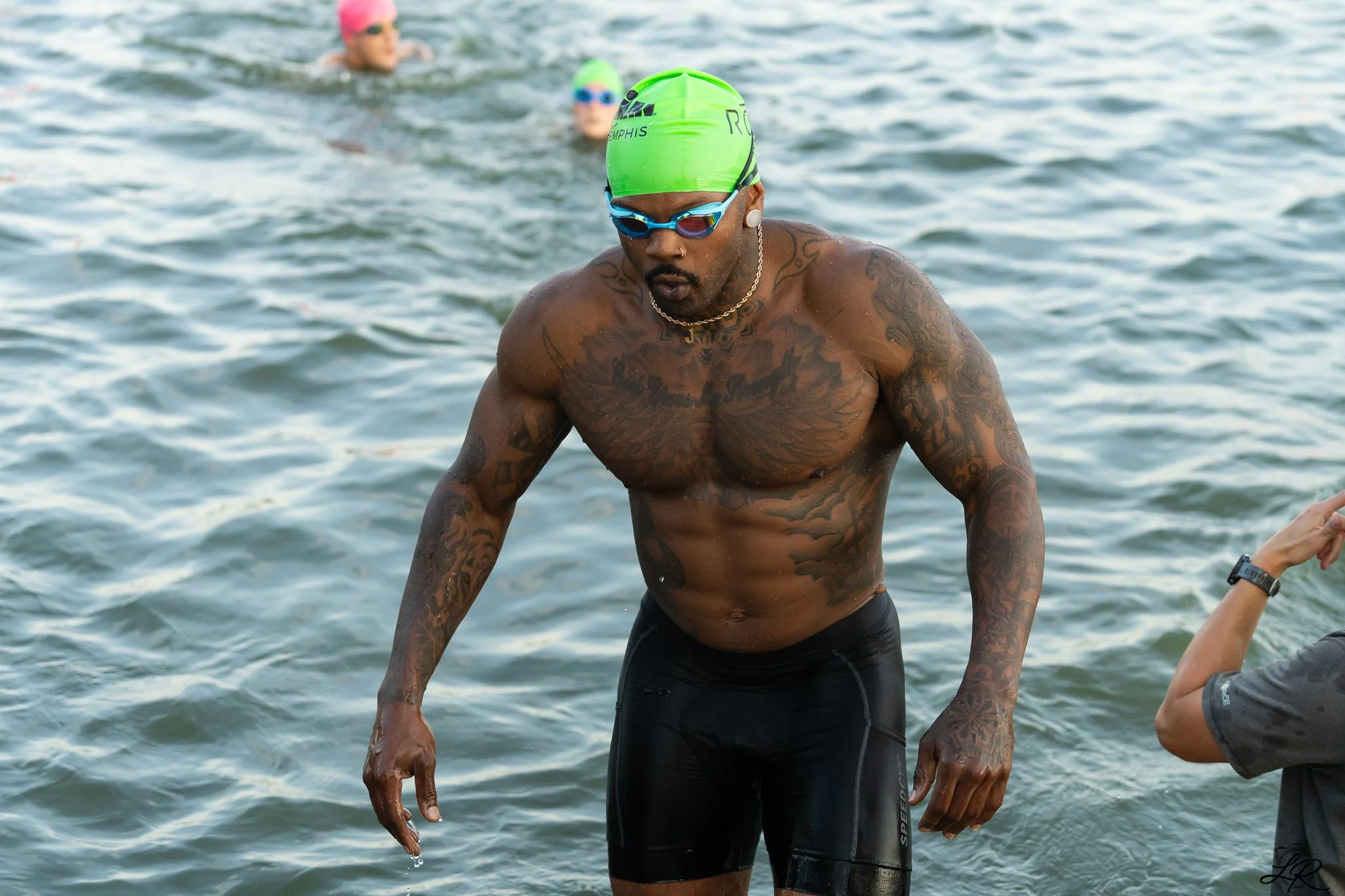 A shirtless man wearing a green swim cap and black swim trunks standing in the water during a swimming event, with two women in the background also swimming.