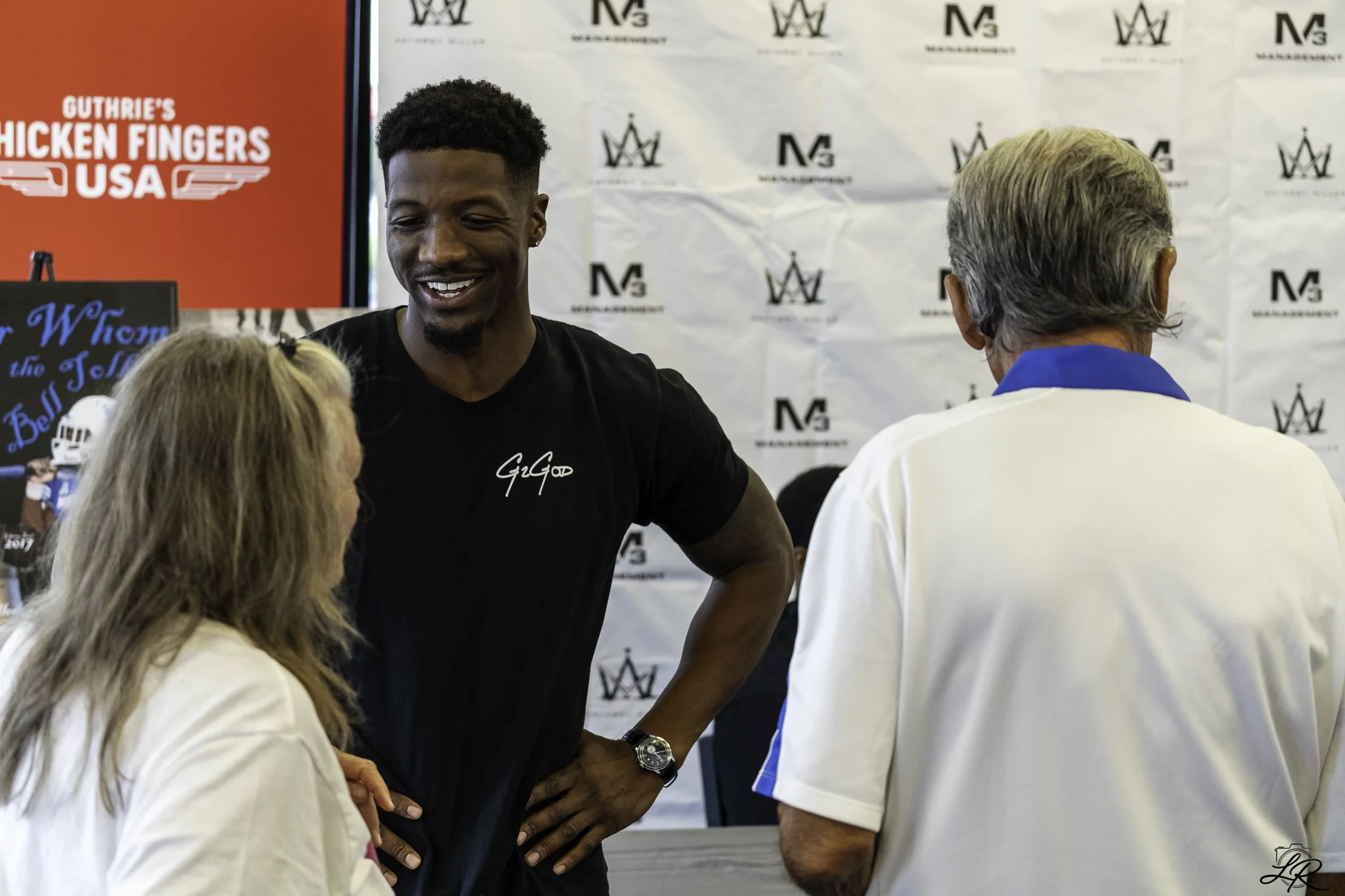 A young man with short curly hair and a goatee smiling and talking to two women at an event with a backdrop that has logos and a sign that reads 'Guthrie's Chicken Fingers USA'.