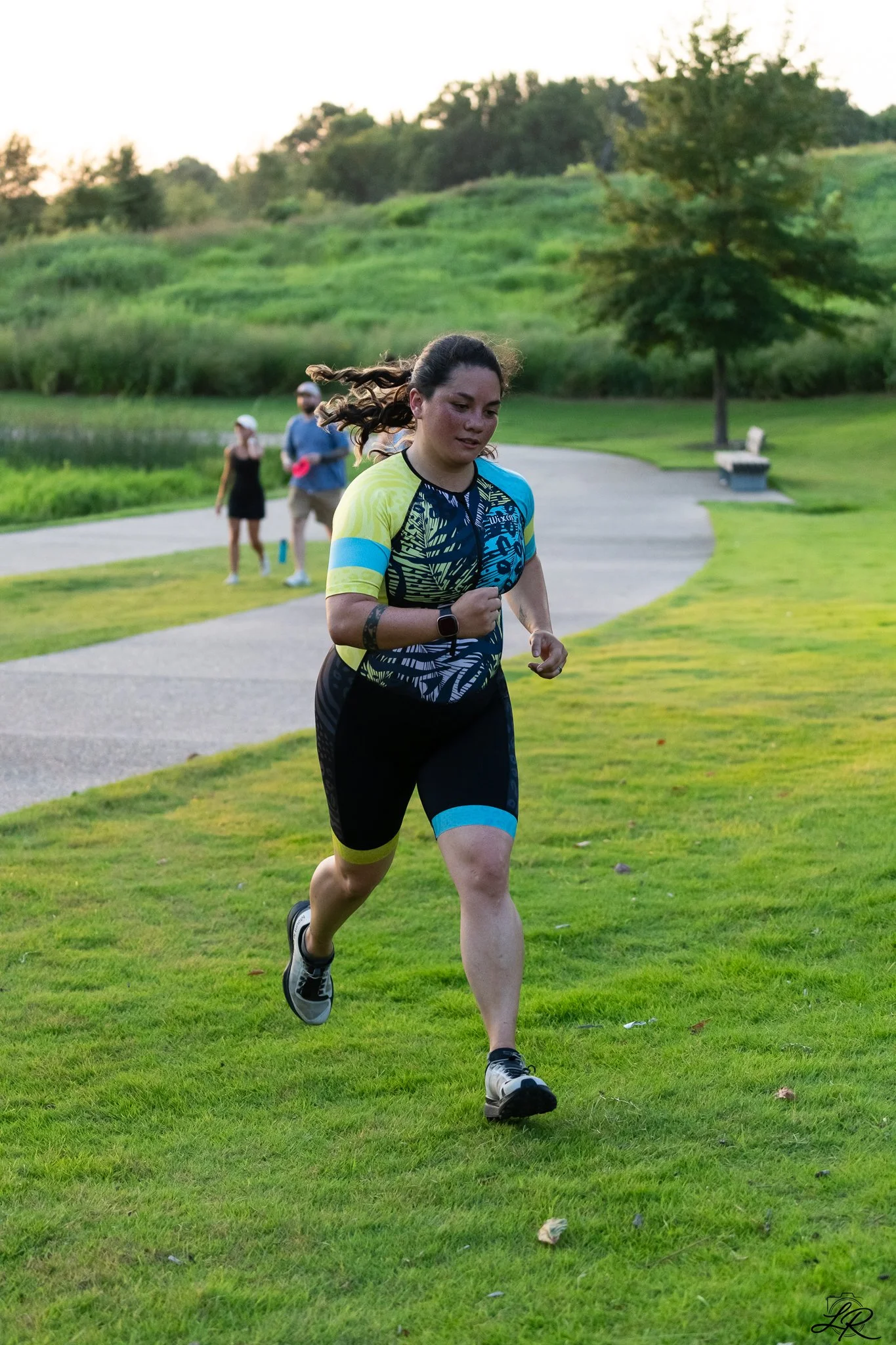 A woman running outdoors on a grassy park path in the evening, with trees and hills in the background and two people standing in the distance.