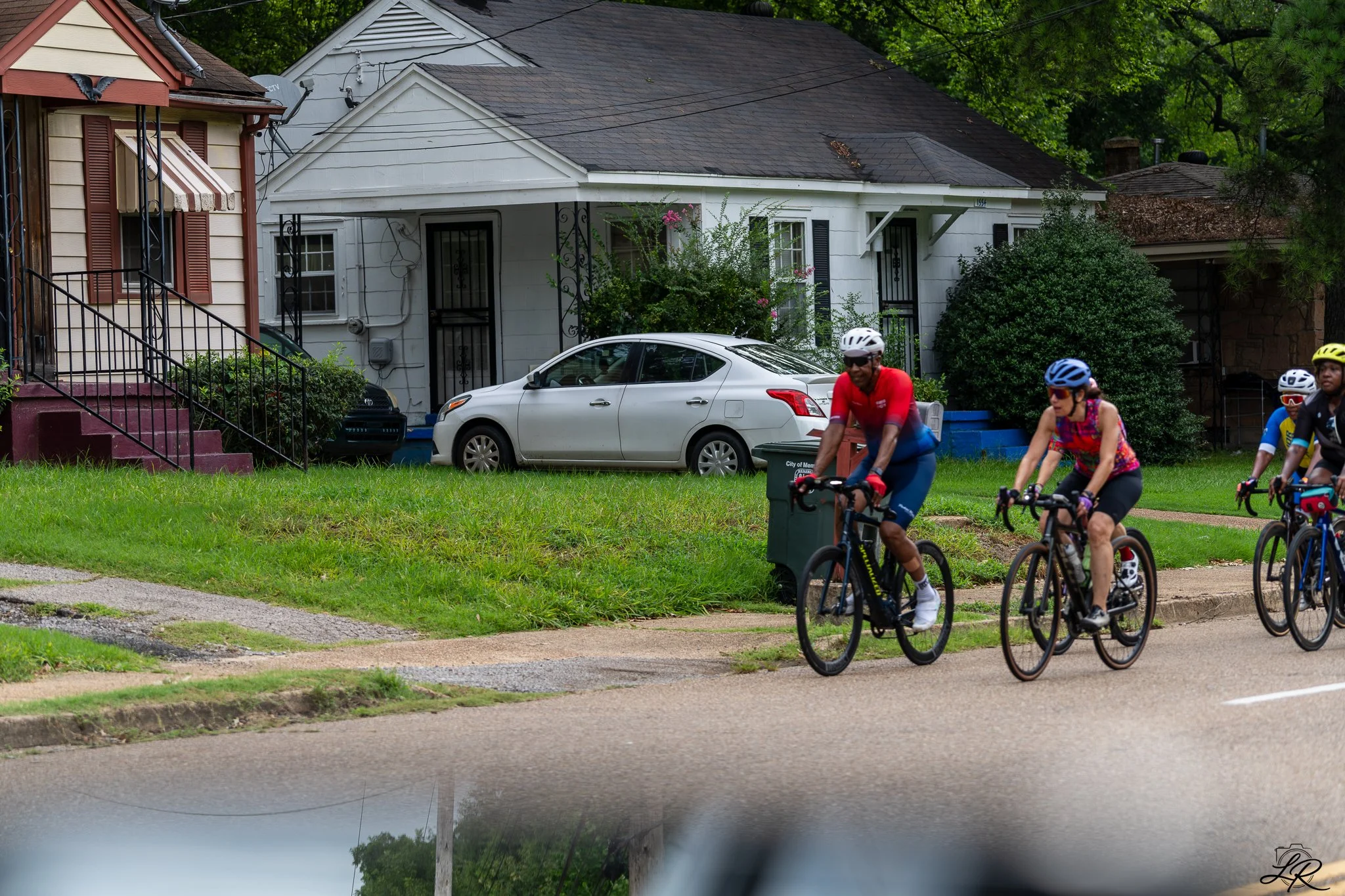A group of people riding bicycles on a sidewalk in front of residential houses with cars parked in the driveway. The scene takes place in a neighborhood with trees and greenery.