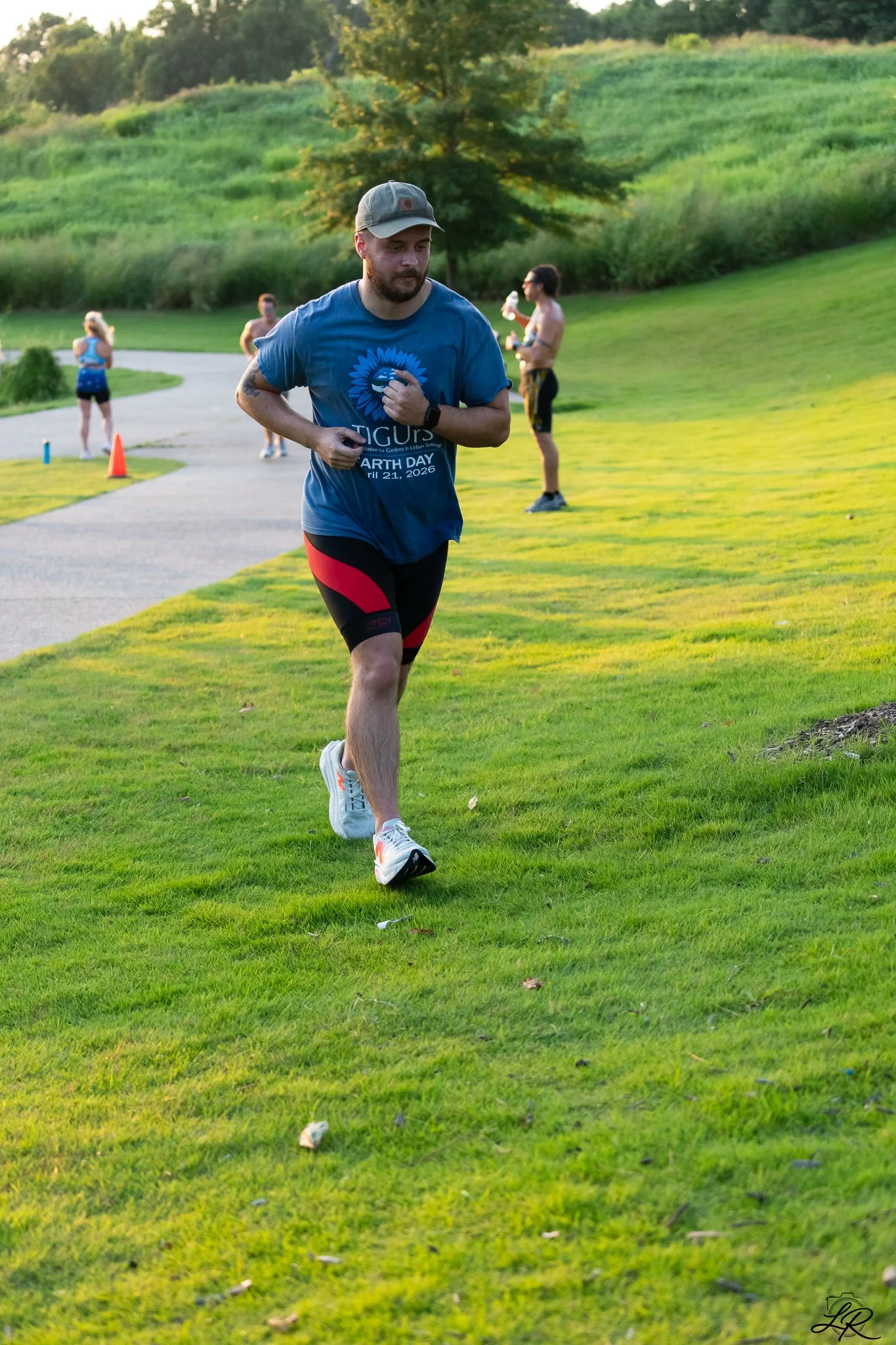 A man running outdoors on a grassy field, wearing a blue T-shirt with a design and black and red shorts, with people in the background and trees in a park or nature setting.