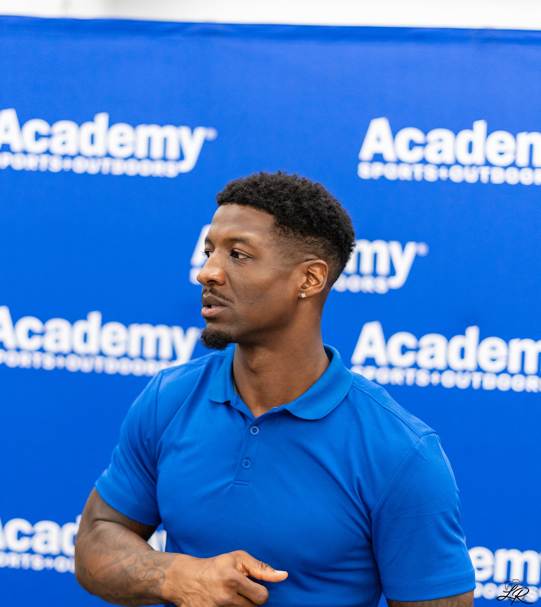 Close-up of a young man with short curly hair, tattoos on his arm, wearing a blue polo shirt, standing in front of a blue backdrop with white text that says 'Academy Sports + Outdoors'.