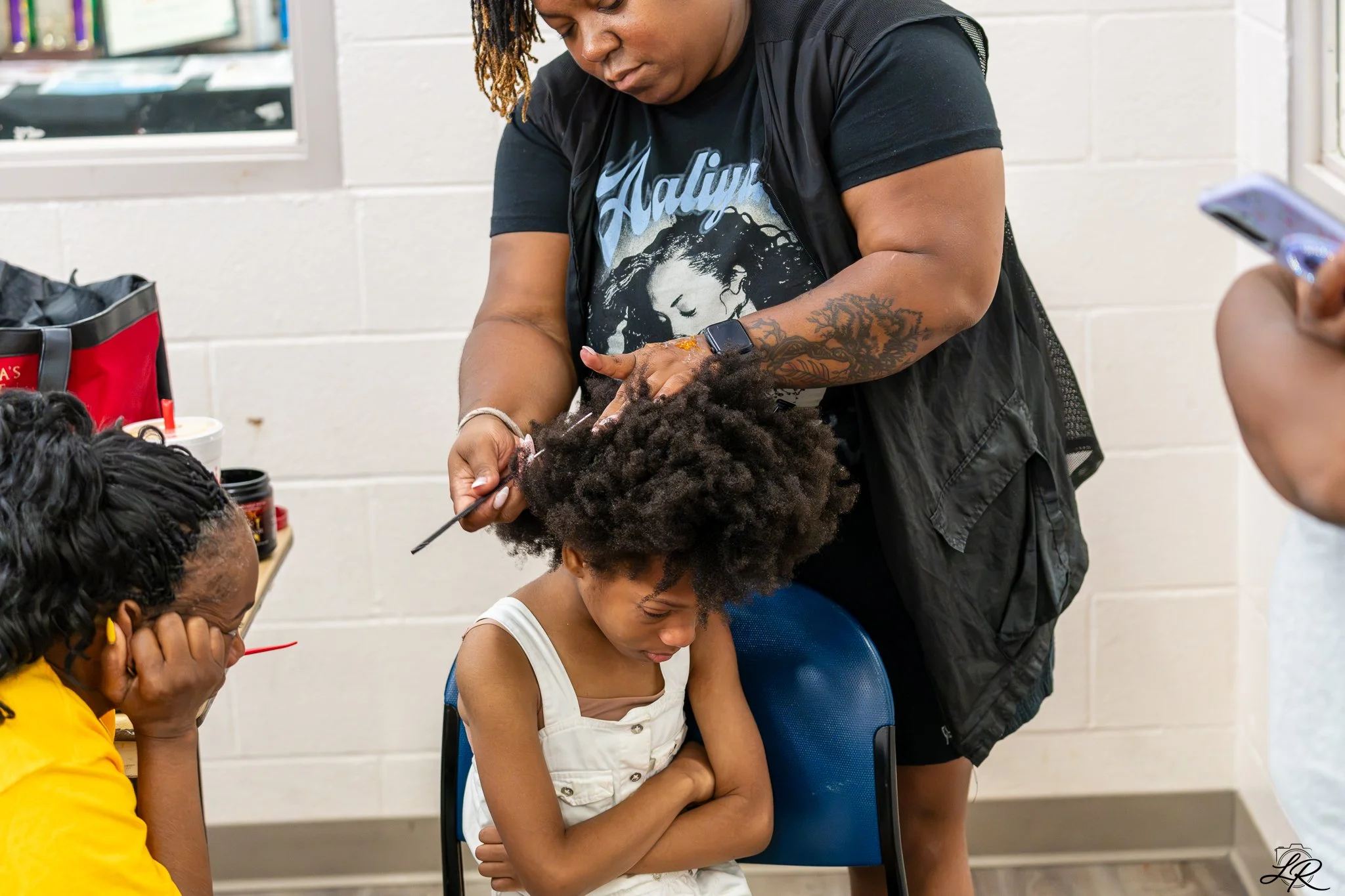 A woman with dreadlocks and tattoos is washing a young girl's hair at a salon. The girl, wearing a white shirt, is sitting with her eyes closed. Other women are seated nearby, observing. There are hair products and tools on the table.