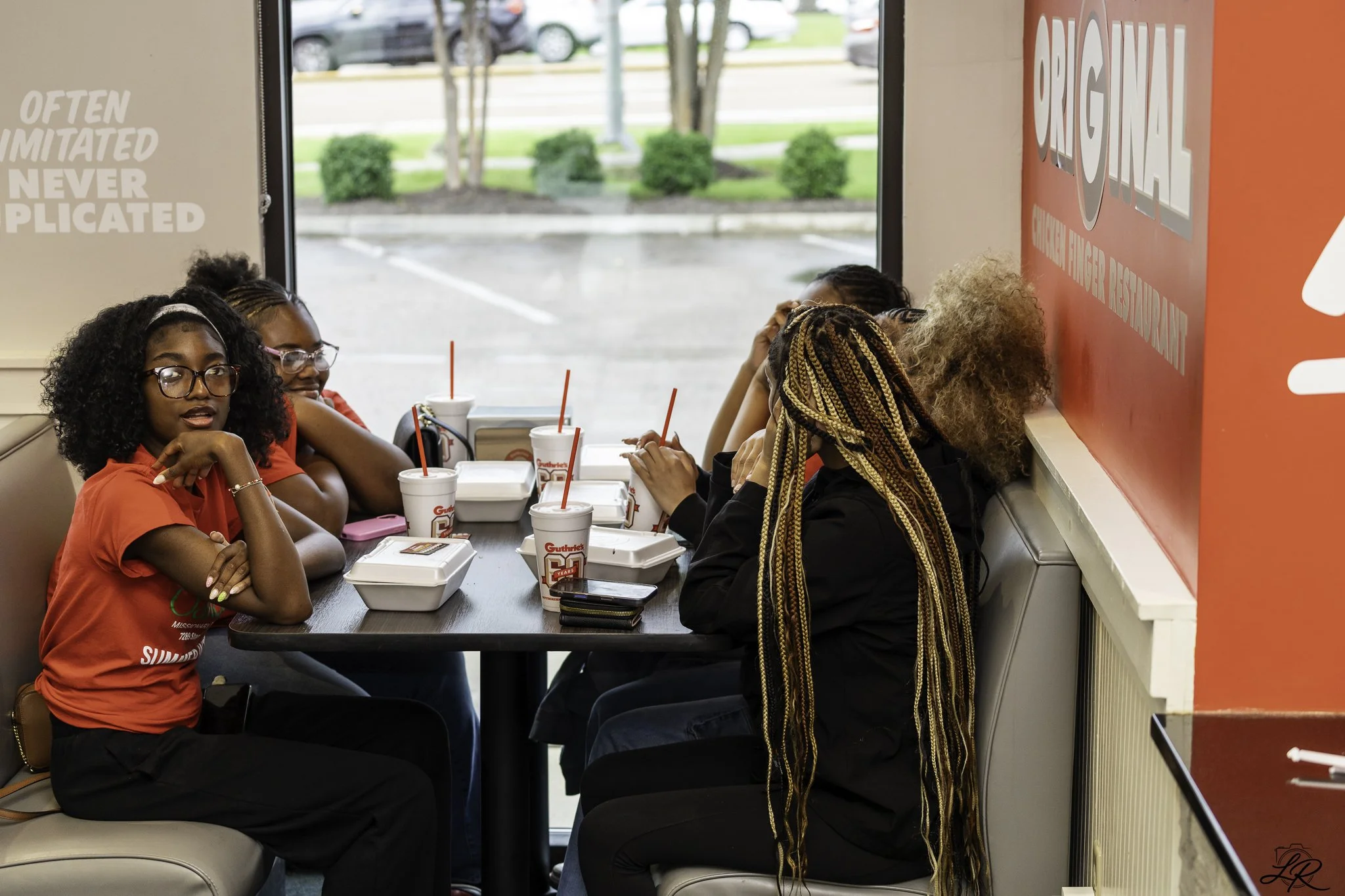 Five women sitting in a fast food restaurant eating. There are drinks with straws on the table, and they appear to be in casual clothing. The restaurant has a window showing parked cars outside.