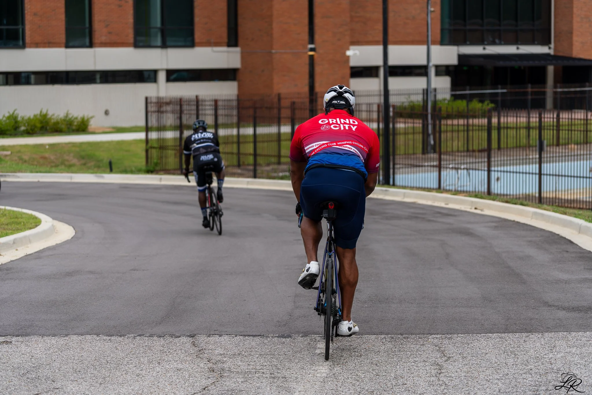 A cyclist in a red jersey with white text and a black and white helmet, riding a bicycle on a curved city street with brick buildings and a fence in the background.