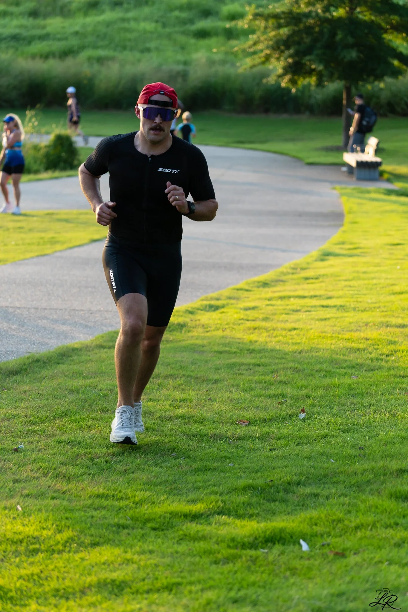 A man in athletic clothing running on a grassy park path with trees and other park visitors in the background during the late afternoon.