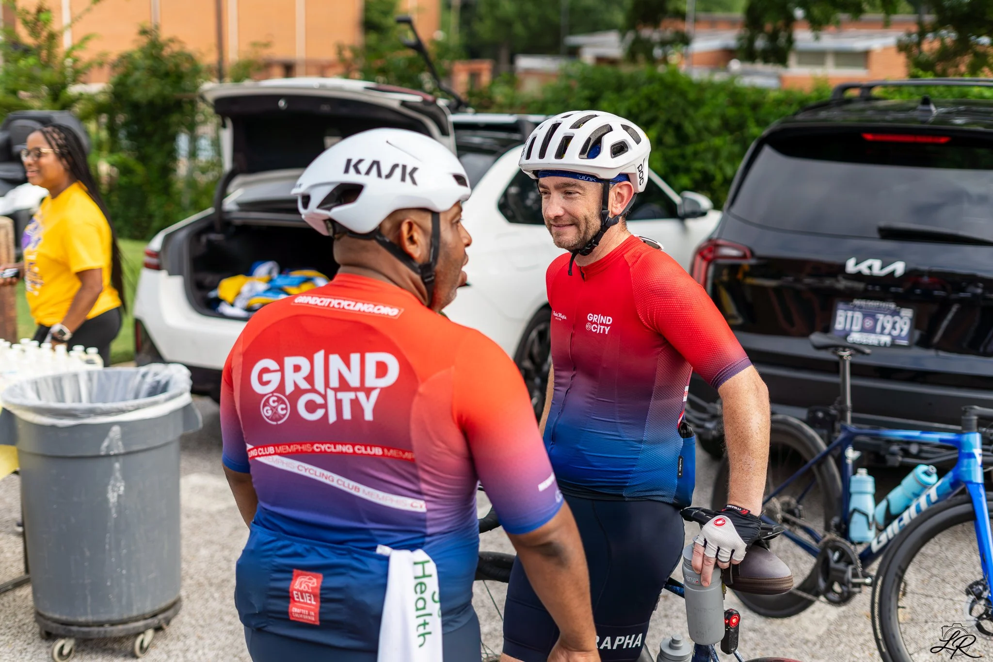 Two male cyclists wearing helmets and athletic gear talking in a parking lot with bikes, cars, and a woman in yellow shirt in the background.