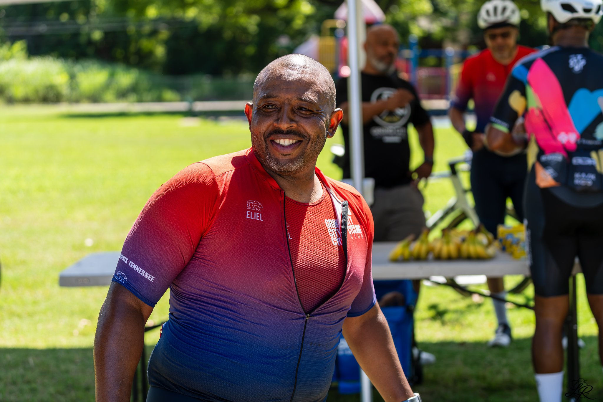 Smiling man in a red and blue athletic shirt at an outdoor event with people and a table in the background.