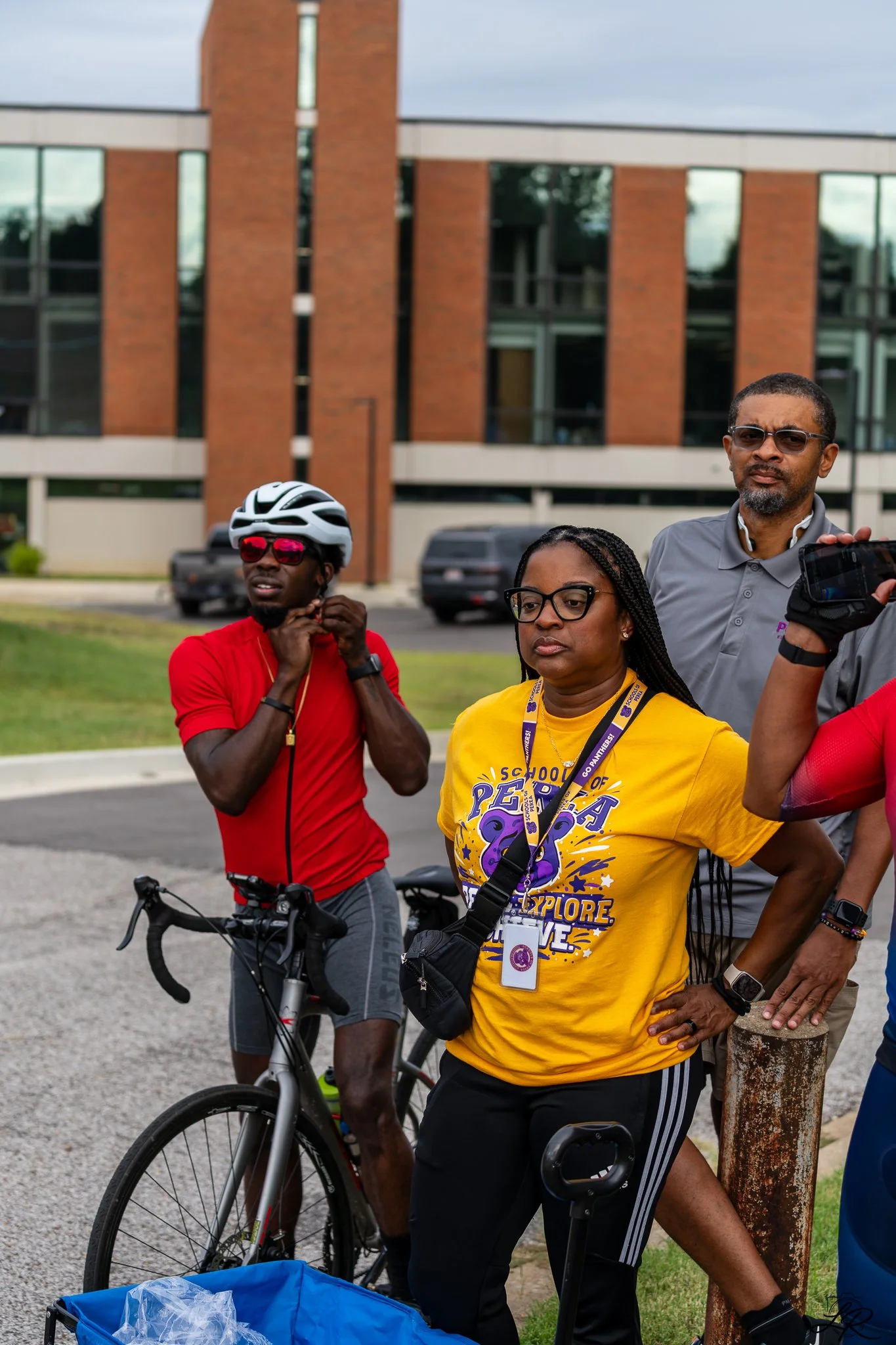 A group of people gathered outdoors in front of a brick building, with one person in a red shirt and bike helmet, a woman in a yellow shirt with a lanyard, and a man in glasses and a gray shirt, standing near a tree.