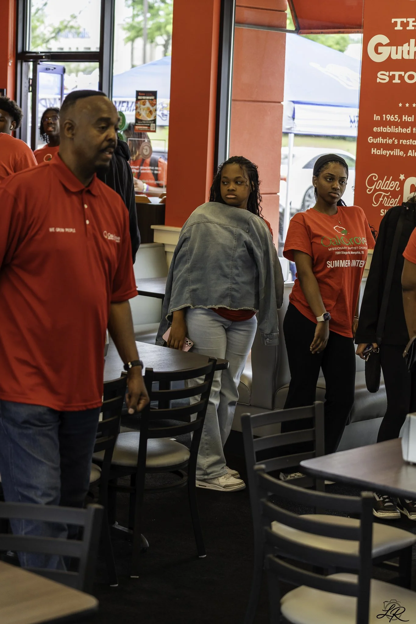 People standing inside a restaurant near the entrance, with some looking to the side and others looking forward. The restaurant has red walls and a large window showing the outside, which includes tents and cars. Some people are wearing red shirts.