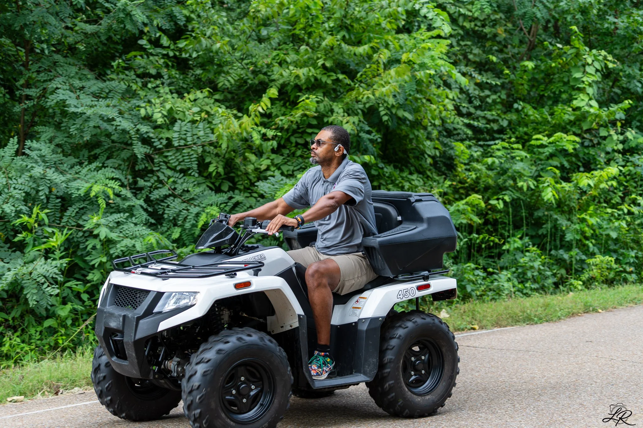 Man riding a white ATV on a path surrounded by lush green foliage, wearing sunglasses, a gray shirt, beige shorts, headphones, and colorful shoes.