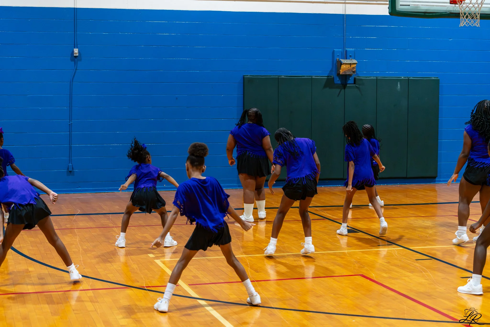 Children and adults in blue outfits practicing a dance or cheerleading routine in a gymnasium with a blue wall and basketball hoop.