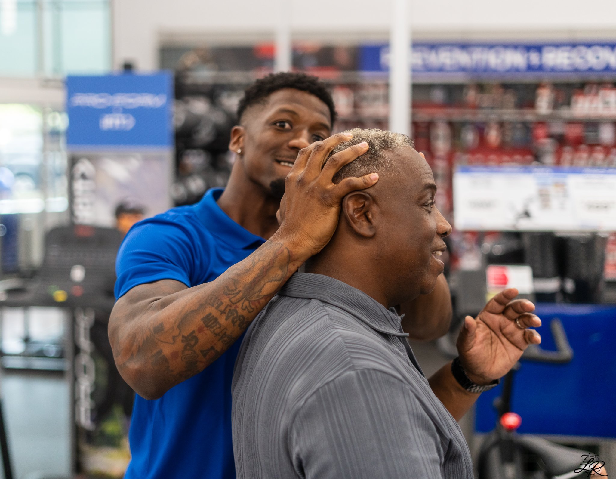 A barber gives a haircut to a customer in a barbershop, with shelves of hair products in the background.